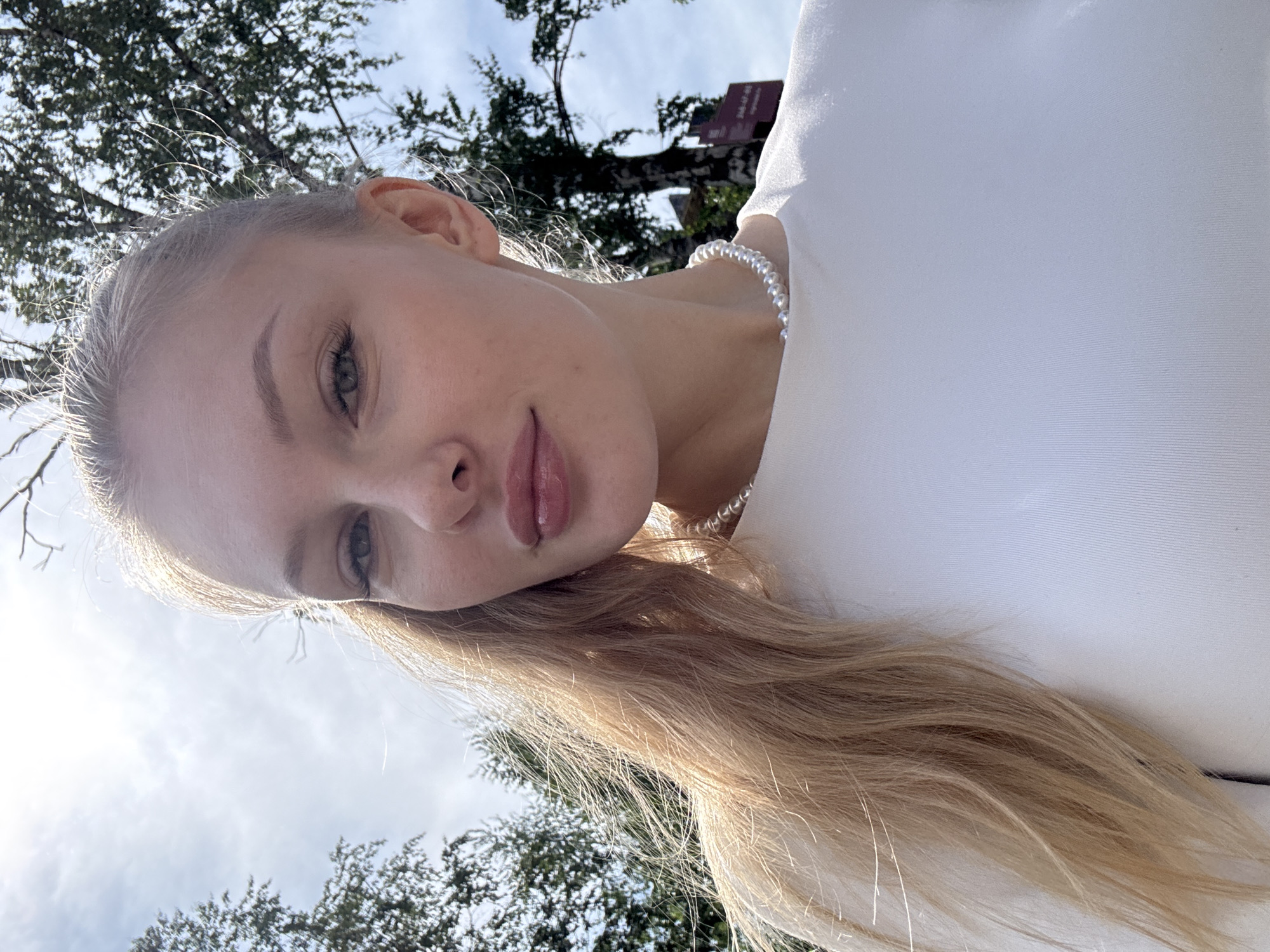 This is a medium close-up outdoor portrait of a young woman with long blonde hair, fair skin, and light-colored eyes, presented rotated 90 degrees to the left. She looks directly at the camera with a soft, slightly smiling expression, her lips gently parted. She wears a white top, and a white pearl necklace is visible around her neck. Bright natural daylight illuminates her face and hair, creating a luminous halo effect on her blonde strands, indicating a daytime shot.
The background shows a bright sky with scattered clouds and the green foliage of trees, suggesting a park or natural outdoor setting. In the upper right background, a dark red sign is partially visible, displaying faint white Cyrillic text that appears to include "300-ЛЕТ" and "Перми." This strongly suggests a commemorative sign, likely referencing the 300th anniversary of the city of Perm, confirming the location as Perm, Russia. The photograph is taken from a low-angle perspective, looking upwards towards the subject and the sky. The woman appears relaxed and calm.