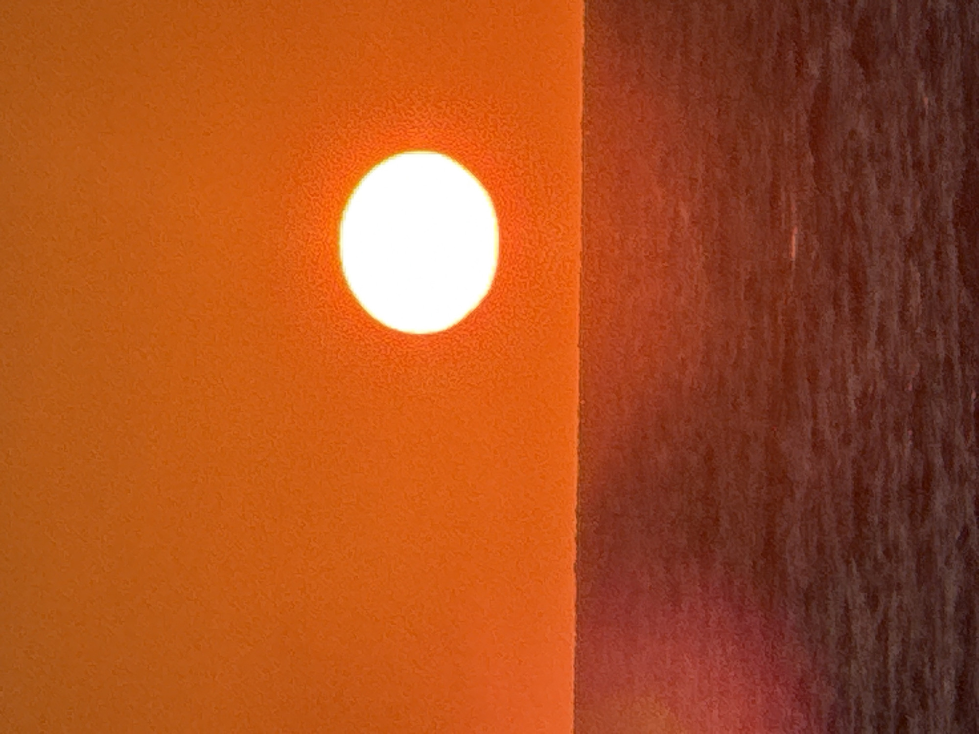 The image features a brilliant, circular white light source, resembling the sun, prominently set against a textured, vibrant orange surface on the left. This intense orange area meets a darker, reddish-brown textured surface on the right along a sharp vertical line, positioned slightly off-center. The orange surface appears brightly illuminated by the sun, suggesting a direct light source. Both surfaces exhibit a subtle, grainy texture.
The scene captures the powerful visual effect of sunlight on architectural or natural elements. The warm, rich color palette, dominated by bright orange and deep reddish-brown, strongly indicates either sunrise or sunset. The sun is visible as a distinct, clear disc, implying generally clear weather conditions. While no specific architectural or landscape features are definitively identifiable, the straight vertical line could represent a building's edge or a wall, and the textured surfaces could be stucco, concrete, or even natural rock formations. The overall impression is minimalist and abstract, focusing on light, color, and texture. Given the location context of Tel-Aviv, Israel, this could depict the sun rising over the sea or setting behind city structures, or vice versa, evoking feelings of warmth and natural spectacle. No text is visible in the image.