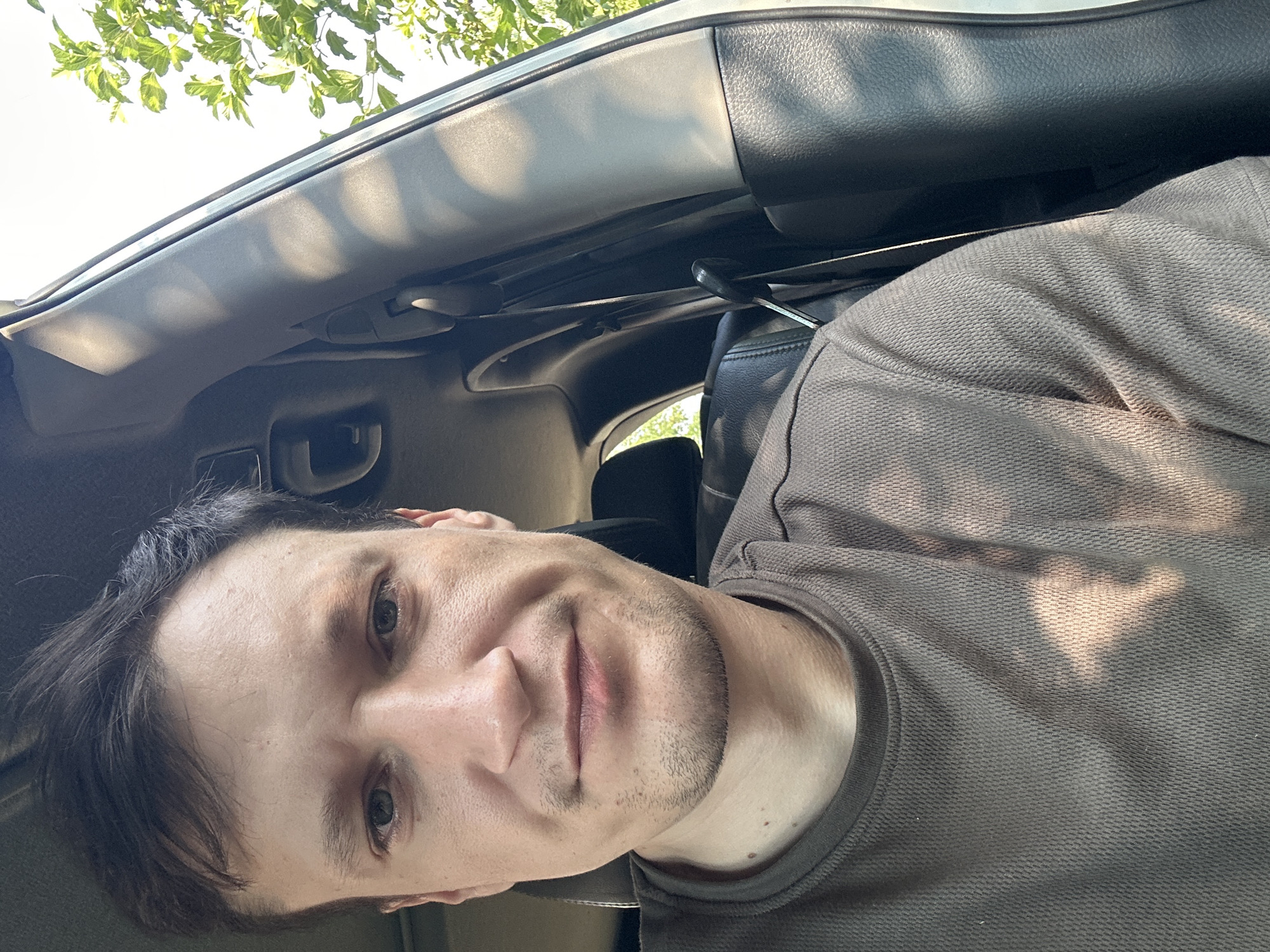 The image is a close-up, selfie-style portrait of a Caucasian man with dark hair, seated inside a car. He is wearing a brown, textured t-shirt. The man's face is turned towards the camera, with a neutral expression. Sunlight casts dappled shadows across his face and the interior of the car. The background shows parts of the car's interior, including a black leather seat, and a glimpse of green foliage through the car window, suggesting an outdoor setting. The specific location of Shatava, Ukraine, is not visually ascertainable from the image itself.