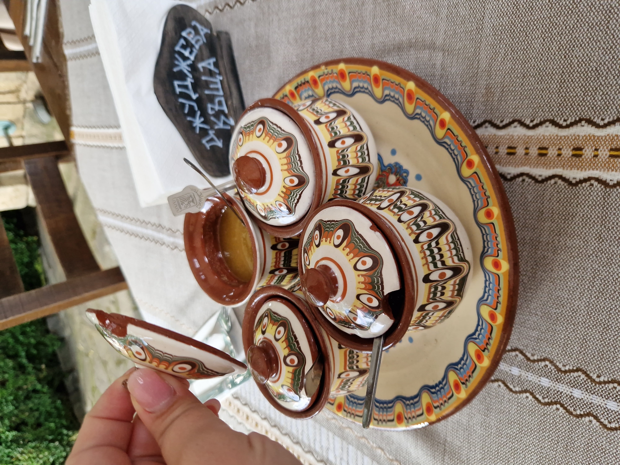 A hand holds a lid of a small ceramic pot, revealing a traditional Bulgarian meal set on a table covered with a patterned cloth. The table is set with four small, lidded ceramic pots, each decorated with colorful, concentric eye-like patterns in shades of brown, yellow, orange, and blue. The pots contain a golden-colored liquid, possibly honey or jam. A larger decorative plate, matching the pots in style, supports the set. To the right of the pots, a dark wooden sign with white lettering in Bulgarian reads "ДЖУДЖЕВА КЪЩА" (Dzhudzheva Kashta), which translates to "Dwarf's House." This suggests the location is a restaurant or establishment in Panagyurishte, Bulgaria, known by this name. In the background, a wooden chair and some greenery are visible, indicating an outdoor or semi-outdoor setting. The overall impression is one of a traditional Bulgarian culinary experience.