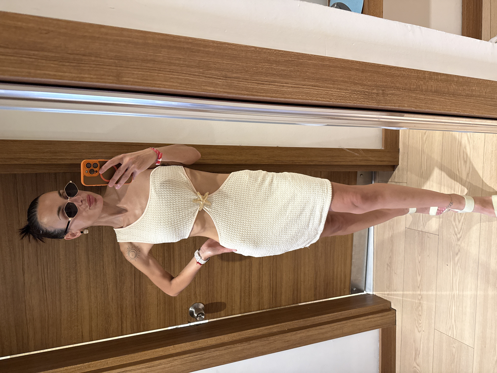 A woman is standing in a room and taking a selfie in a mirror. She is wearing a white, one-shoulder, cutout mini dress and white heeled sandals. She has sunglasses on and her hair is pulled up in a bun. The room has wooden doors and a wooden floor. The overall aesthetic is modern and minimalist. This image was taken in Aksu, Turkey.