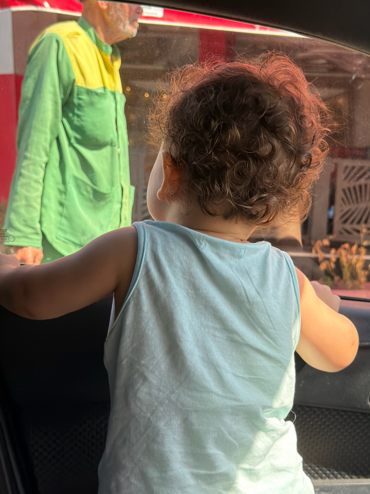 An infant, viewed from behind and from the shoulders up, looks out of a car window. The infant's curly brown hair is illuminated by warm light. They are wearing a light blue sleeveless shirt. In the background, a man wearing a green and yellow shirt is visible through the window, partially obscured. The scene appears to be in a city, possibly Sharm El Sheikh, Egypt, indicated by the architectural details in the background and the presence of a car.