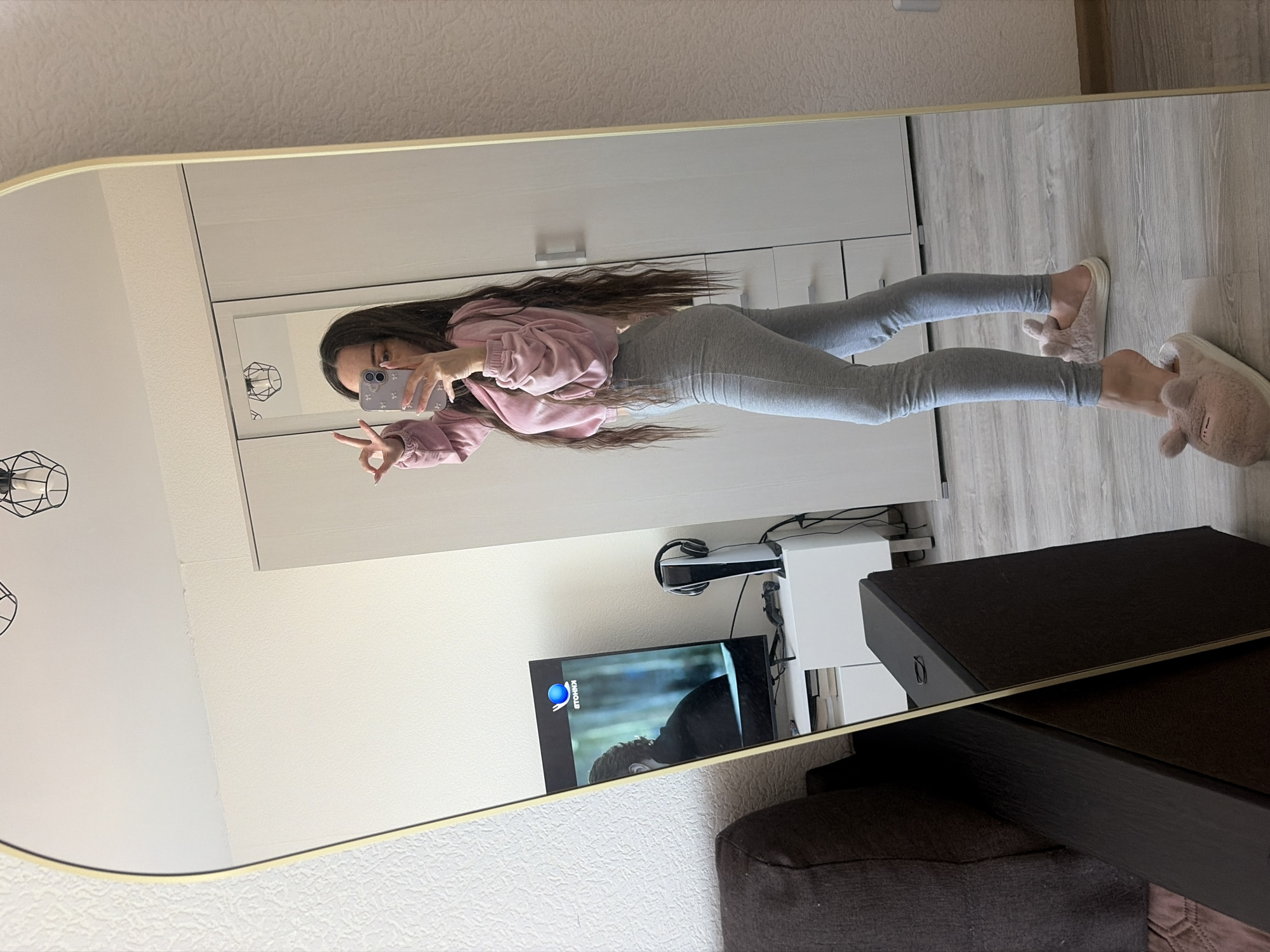 A young woman in grey leggings and a pink hoodie is standing in front of a mirror, holding up her phone to take a selfie. She is making a peace sign with her left hand. She is also wearing fluffy, animal-shaped slippers. The mirror reflects a room with white walls, a large white wardrobe, a TV on a stand next to a Playstation 5, and a dark-colored dresser or table. The floor is light-colored wood laminate. A ceiling fixture with a black wireframe design is visible in the reflection.
It is not possible to determine the location within the city of Saint Petersburg, Russia, based on this image.