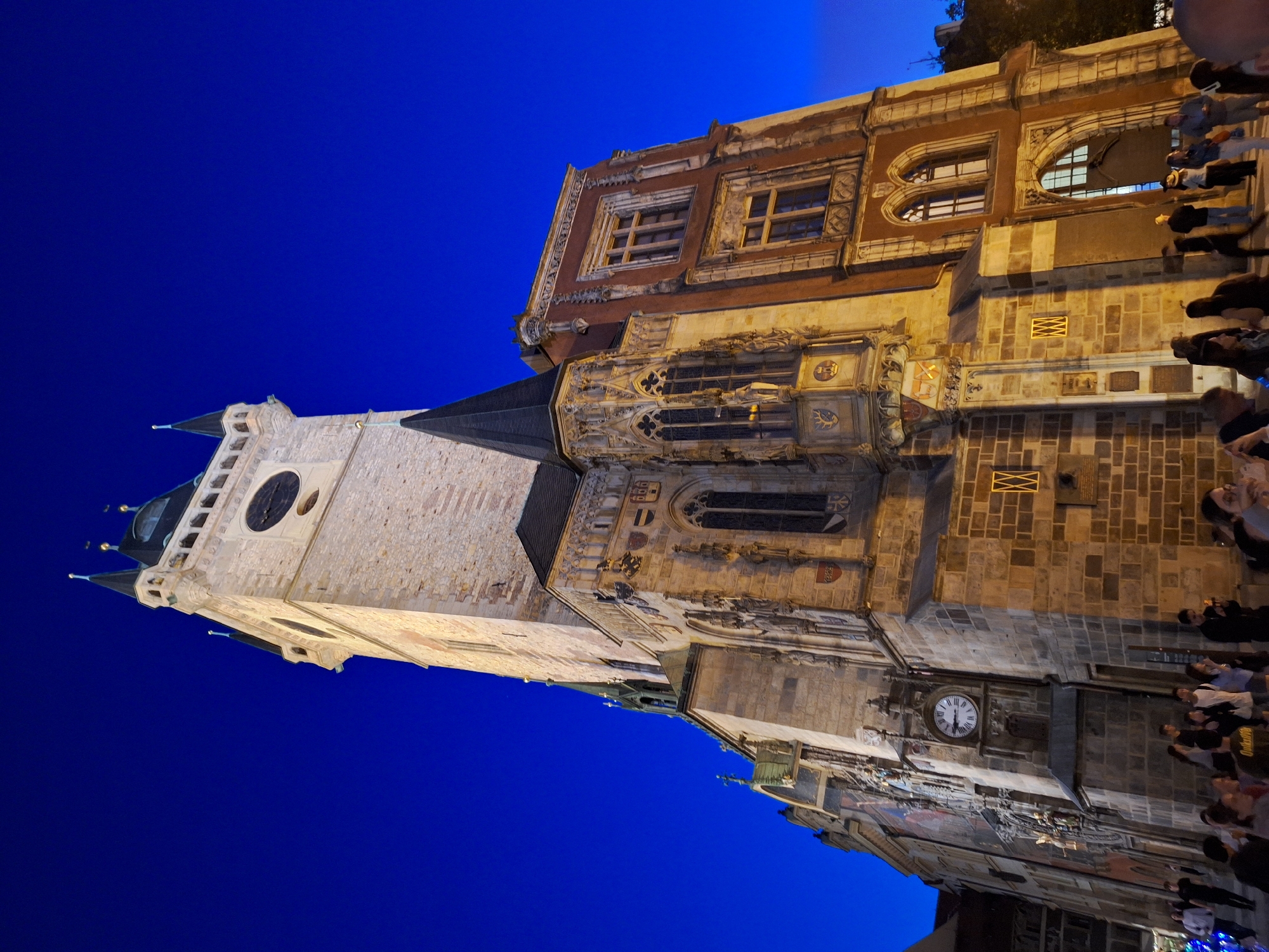 The image shows a low-angle, nighttime view of the Old Town Hall Tower in Prague, Czechia. The tower, constructed of stone and featuring a clock and spires, is illuminated by artificial lighting, creating a warm glow against the deep blue night sky. The tower is an intricate Gothic structure with ornate carvings and decorative elements. Attached to the tower is a red-bricked building with arched windows. In the foreground, a crowd of people, indistinct in the low light, are gathered in the square, some looking at the tower, others conversing. The overall scene depicts a popular tourist location at night.