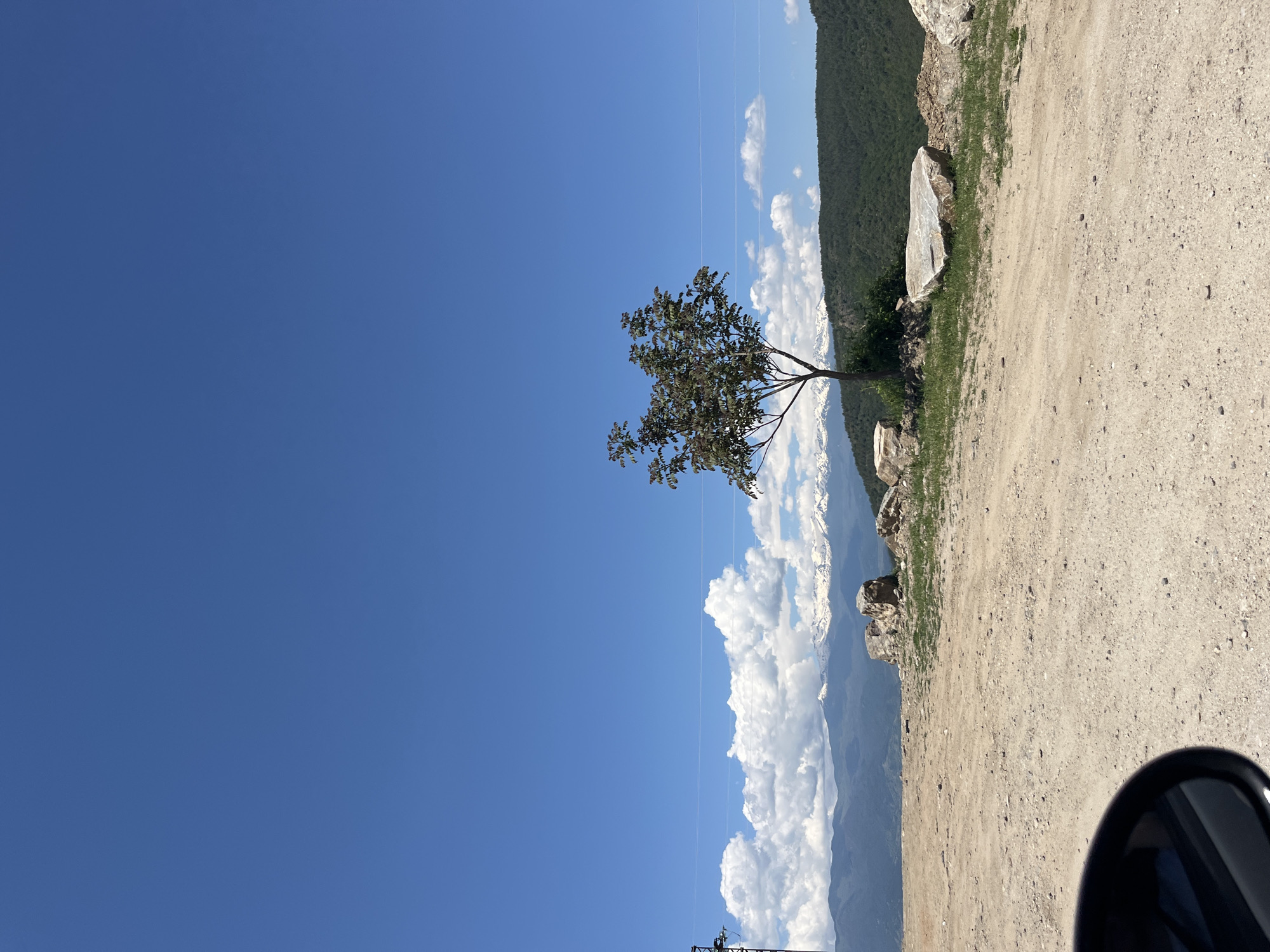 The image shows a landscape with a dirt road on the right side and a clear blue sky on the left. A lone tree stands between the road and a mountain range partially obscured by large white clouds. Behind the mountain, a green hillside is visible. The bottom right corner shows a side mirror. The image appears to be taken in a natural setting. It is not possible to determine the specific location in Leshten, Bulgaria.