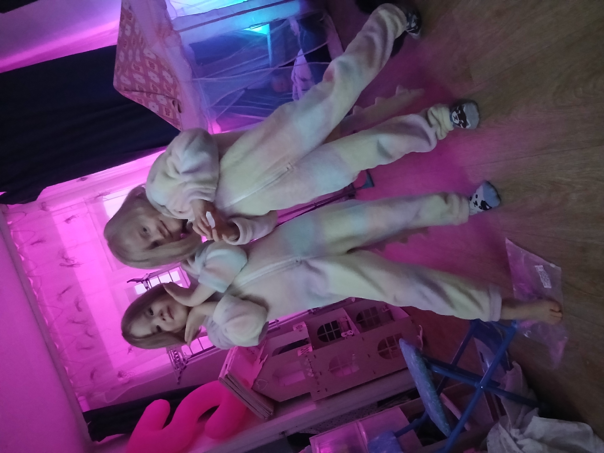 Two children, one with blonde hair and the other with brown hair, lie on a wooden floor, both wearing pale-colored animal-themed onesies. They are indoors, near a pink inflatable object and a dollhouse. The environment is illuminated with pink and blue lighting. A chair and some plastic containers are also visible. The location is Chaikovsky, Russia.