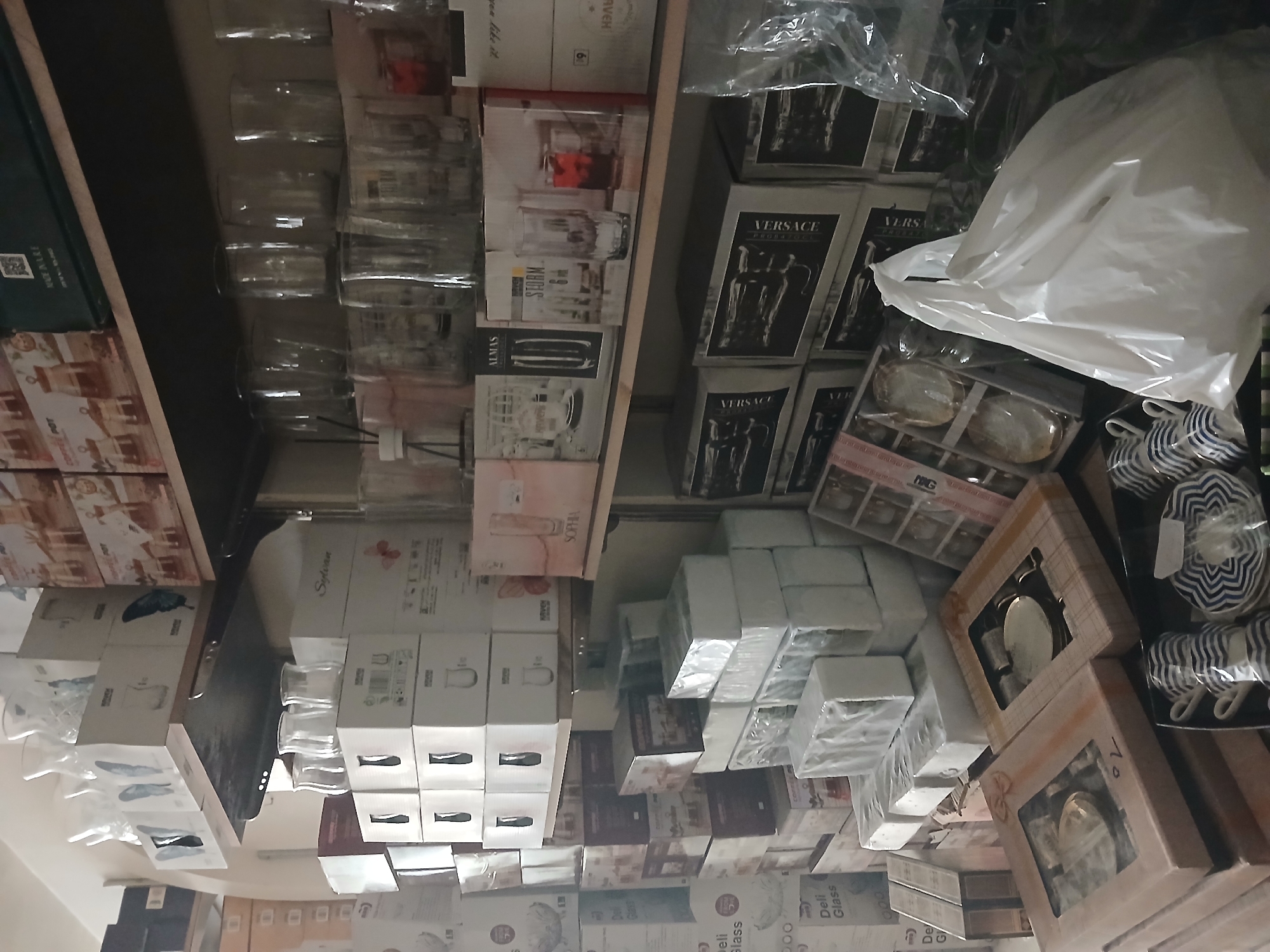 This is an indoor, eye-level, medium shot of a shelf in a store displaying glassware and boxed dinnerware. The shelves are packed with various items, predominantly boxes of glassware and what appear to be tea or coffee sets. Some of the boxes are open, revealing the contents, while others are sealed. On the top shelf, there are several clear glass vases and decorative items. Below, there are stacks of white boxes with images of glassware on them, and some boxes are branded with "Sylvan" and "Sophia". Further down, there are boxes with darker packaging, some featuring intricate designs, with one box labeled "Versace". In the foreground, on a lower shelf, there are two boxed sets of what appear to be tea or coffee cups and saucers. Next to these, in a black tray, are several blue and white striped cups and saucers, some of which have plastic wrapping. A large white plastic bag is prominently placed in the foreground. The lighting is moderate, and the overall impression is of a well-stocked retail environment. The store is located in the city of Rukn ad-Din Municipality, Syria.