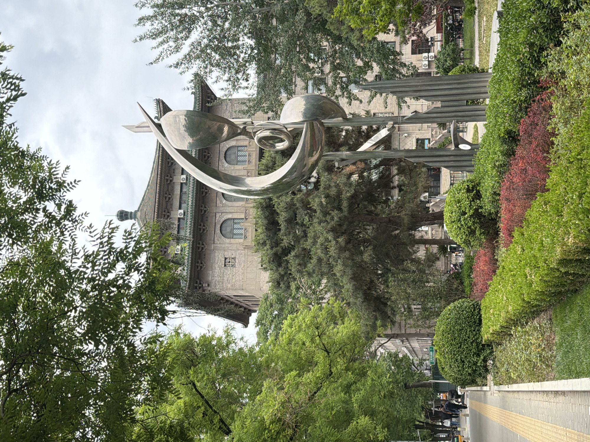 The image captures an outdoor scene in Xicheng District, China. A modern metal sculpture stands prominently in the foreground, featuring curved, shiny surfaces. Behind the sculpture, an older building with traditional Chinese architectural elements, like a multi-tiered roof, decorative eaves, and arched windows, can be seen. The building appears to be made of stone or brick.
The surrounding environment includes various trees and bushes, some neatly trimmed into hedges. People can be seen walking on a paved path in the lower part of the image. The weather is overcast.