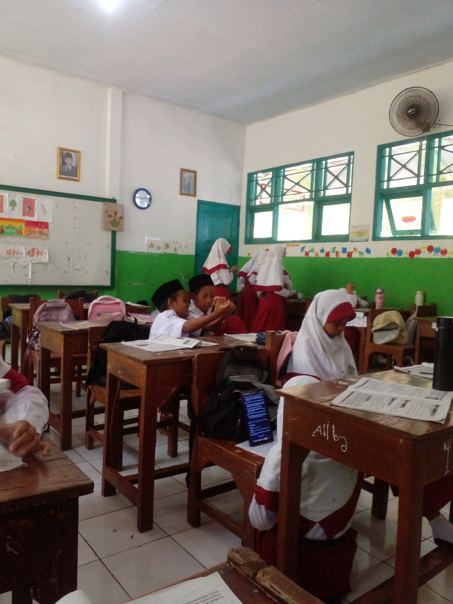 This is a medium-shot, eye-level interior photograph of a classroom in Banyuputih, Indonesia, likely a school. The room is brightly lit, with a white ceiling and cream-colored walls. A large green band runs along the lower portion of the walls, decorated with colorful drawings of small figures. Two windows with green frames and white bars are visible along the back wall, letting in natural light. A ceiling fan is mounted on the upper right wall. A portrait of a man in a suit and hat hangs on the left wall, alongside a framed picture of a landscape. A circular blue clock is also present on the wall.
The classroom is furnished with rows of wooden desks and chairs. Several students, appearing to be of primary school age, are seated at the desks. They are wearing white shirts and maroon skirts or shorts, and many are wearing white headscarves, suggesting a Muslim school. In the foreground, a student in a white headscarf is seated at a desk, with their back to the camera, looking down at papers. To the left, another student is partially visible, with their hand resting on a desk. In the middle ground, two male students are seated at a desk, one of whom is holding a smartphone. Other students are visible in the background, some standing and some seated. Backpacks and books are scattered on the desks and floor. A stack of papers is on the desk in the foreground. The floor is tiled with white squares.