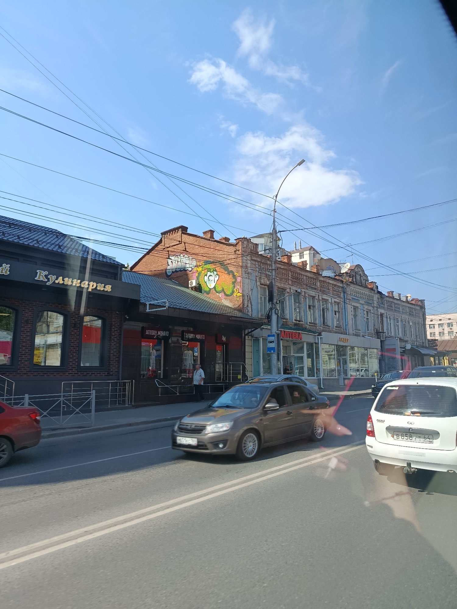 The image shows a street scene in Saratov, Russia, with several buildings lining the road and cars driving on the asphalt. On the left side of the street, a building with the sign "Кулинария" (Culinary) is visible, featuring a dark facade with large windows. Next to it is another building with the "ANTONY MORATO" signage. Further down the street, an "APTEKA" (Pharmacy) sign is prominent on a building with a pale blue facade, exhibiting ornate architectural details. The sky above is clear blue with scattered white clouds. Several cars, including a white Lada Kalina, are on the road, and a person is standing on the sidewalk in front of one of the shops. The street appears to be a commercial area with shops and businesses.