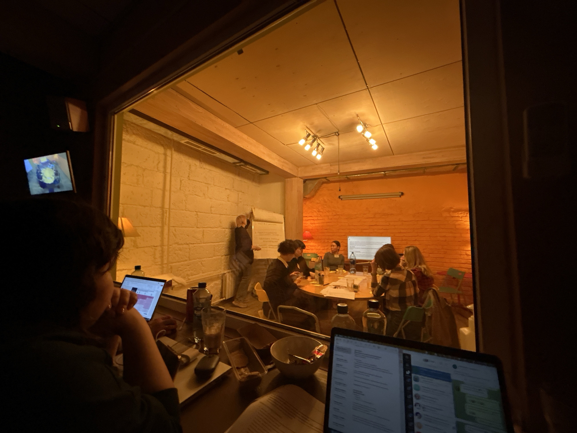 The image depicts a room with a meeting taking place. A group of people is seated around a table in the center of the room, seemingly engaged in a discussion or presentation. One person is standing near a whiteboard, possibly leading the meeting or presenting information. The room has an orange accent wall and an exposed brick wall. In the foreground, outside the room, a person is sitting at a desk working on two laptops.
Without additional information, it is impossible to determine whether the image was taken in Moscow, Russia.