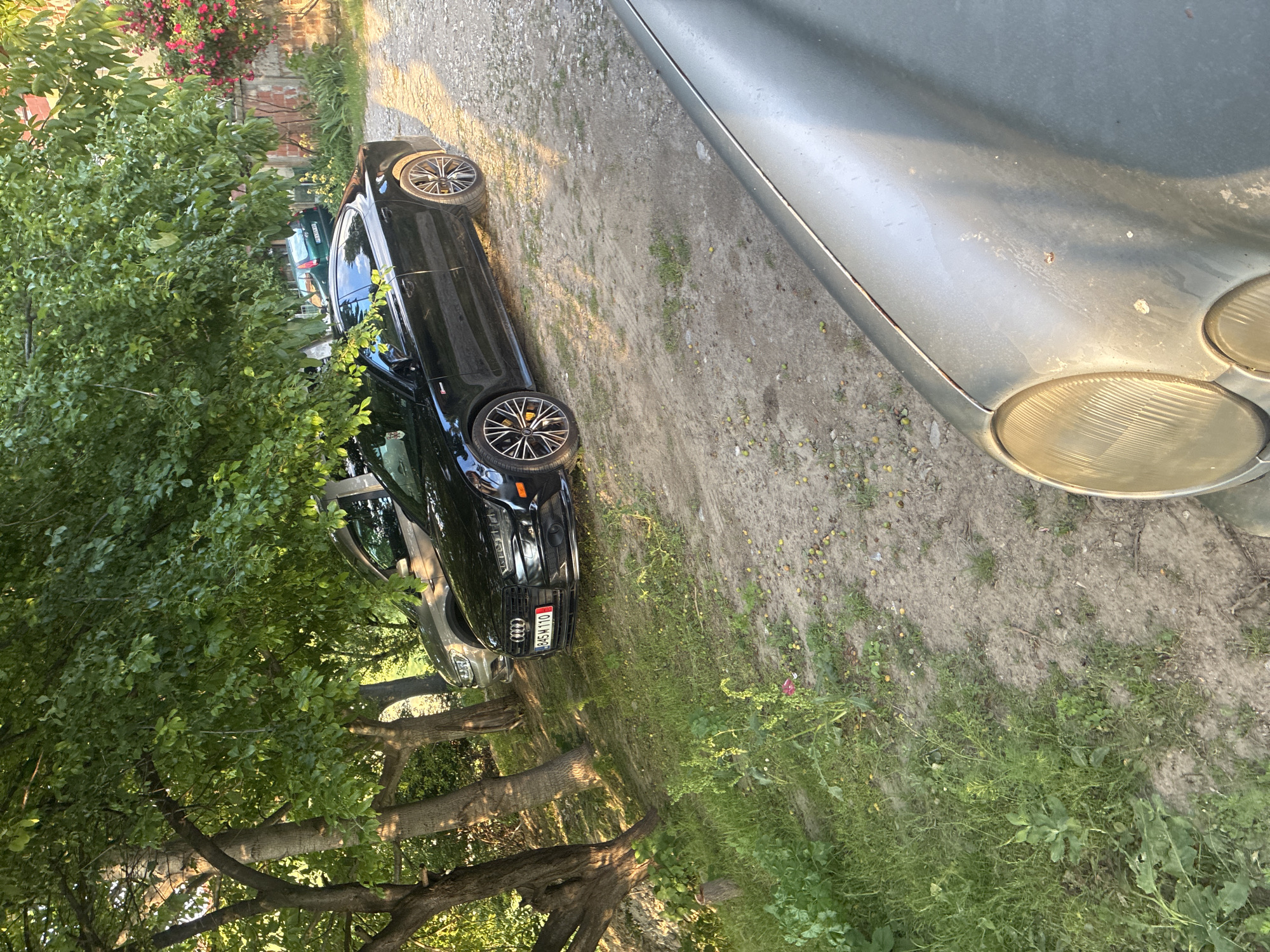 The image shows a partially obscured black Audi sedan parked on a dirt and gravel surface. Part of another car, possibly a silver Mercedes-Benz, is visible in the top right corner. The Audi has an Austro-Hungarian license plate number, B454M-110. Trees and foliage are visible to the left of the cars. The location appears to be outdoors and is likely a parking area or driveway. No people are visible. The city and country are unknown.