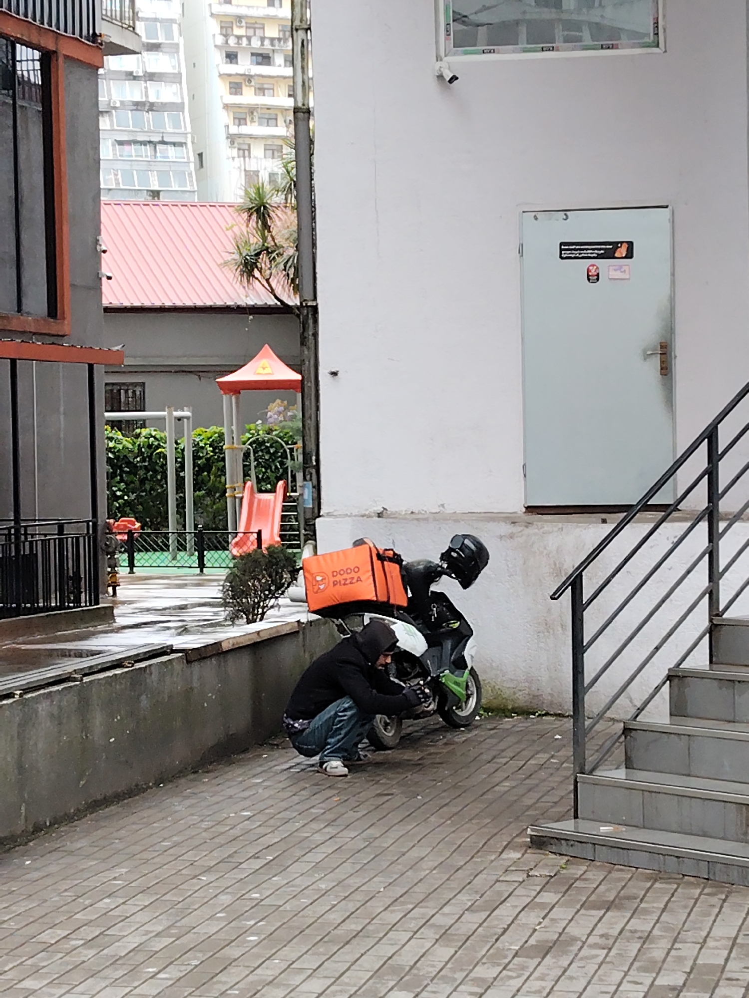 A delivery worker is squatting next to a green scooter parked outside a white building in Batumi, Georgia. The scooter has a black helmet and an orange "Dodo Pizza" delivery box attached. The man is wearing a black hoodie, jeans, and white shoes. In the background, there's a playground with a slide and a multi-story apartment building. The weather appears overcast. The scene is on a paved walkway, with stairs to the right. A security camera is present on the white building above the door.