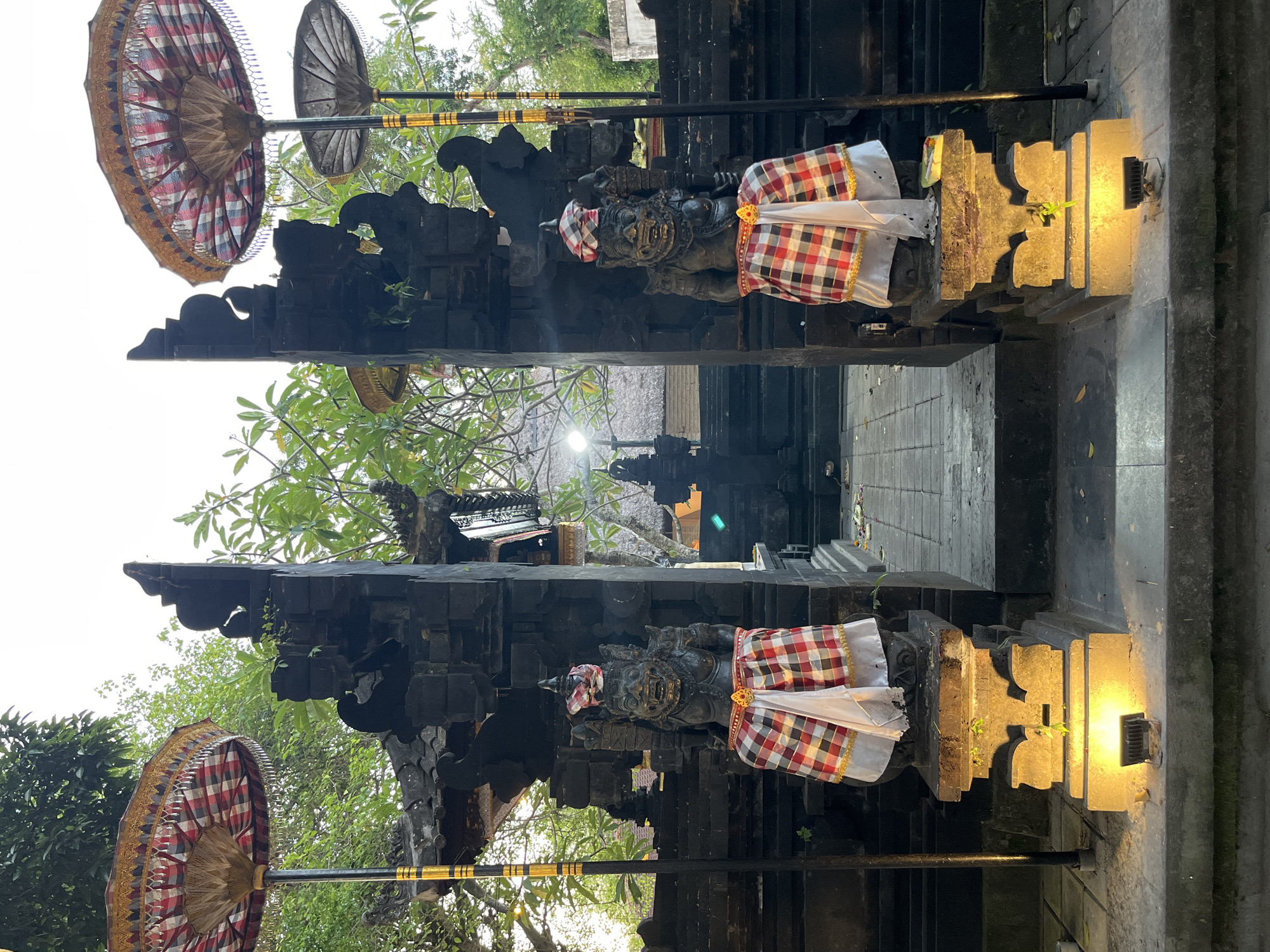 This is a vertical, eye-level, medium shot of a traditional Balinese temple entrance in Kuta Selatan, Indonesia. The entrance is flanked by two dark stone statues of mythological guardians, each wearing a checkered sarong and a decorative headpiece. The statues are illuminated from below by warm yellow lights. In front of the statues are two large, ornate umbrellas with red and white checkered patterns and gold trim. The umbrellas are supported by tall poles with gold and black accents. Behind the entrance, lush green trees and foliage are visible, and the sky is bright white, suggesting it is either early morning or late afternoon. The architecture of the temple is characterized by dark, rough-hewn stone, with tiered roofs and intricate carvings. The ground is paved with stone tiles. There are no people visible in the image.
