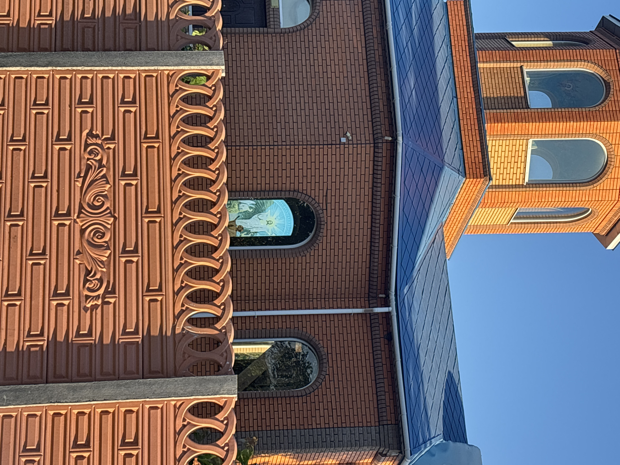 The image presents a daytime architectural view featuring several distinct building sections under a clear blue sky. The photo is rotated, showing the buildings vertically.
On the left, there is a wall adorned with elaborate terracotta-colored panels. These panels feature a pattern of raised vertical rectangles, interspersed with decorative relief sculptures of flowing, organic designs and scrollwork. A prominent border runs along the top and right edge of this section, composed of intricate interlocking circular motifs. The strong, low-angle sunlight casts sharp shadows, emphasizing the texture and three-dimensionality of these decorative elements.
Adjacent to this ornate wall, a larger building constructed of red brick is visible. It features several arched windows. One window in the mid-section contains what appears to be a stained-glass artwork depicting a luminous figure, possibly a religious icon, in shades of white and blue. Another arched window below it reflects trees or greenery. Further right, a multi-story tower-like structure, also built from red brick, rises into the sky, featuring more arched windows.
A prominent blue structure, resembling an awning or a roof extension with a grid pattern, extends diagonally across the mid-ground, connecting to the main brick building. White drainpipes are visible on the brick wall near this blue structure.
The scene is brightly lit by natural sunlight, evidenced by the vivid blue sky and the distinct shadows cast by the architectural features. No visible text is present in the image.