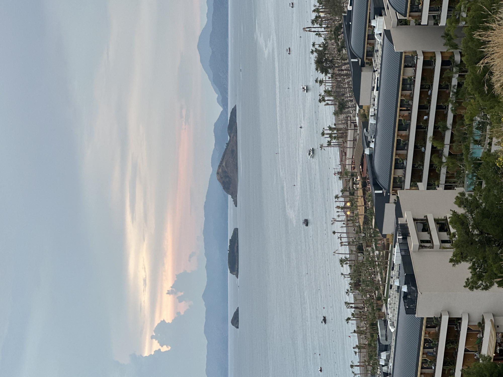 The image depicts a coastal scene in Fethiye, Turkey. The view is taken from an elevated position, possibly a building overlooking the sea. The sky is partially cloudy, with hues of orange and gray near the horizon. The sea is calm, and several small boats are scattered across the water. In the distance, mountain ranges are visible. The coastline is lined with palm trees and buildings that appear to be part of a resort or residential complex. The architecture of the buildings is modern, with multiple balconies and terraces. Some vegetation is visible near the building from which the photo was taken.
