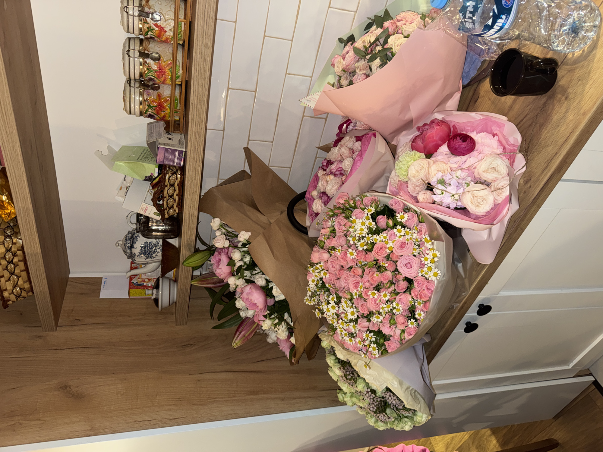 The image shows a collection of flower bouquets on a wooden surface, possibly a counter or shelf, situated next to a white-tiled wall and a shelf displaying decorative items. Five distinct bouquets are visible, each wrapped in different colored paper (pink, brown, purple). The flowers consist of roses, daisies, and possibly lilies. In the background, bottles of water and a black mug are present, suggesting an indoor setting, perhaps a home or a flower shop. The context is Unknown, Unknown.