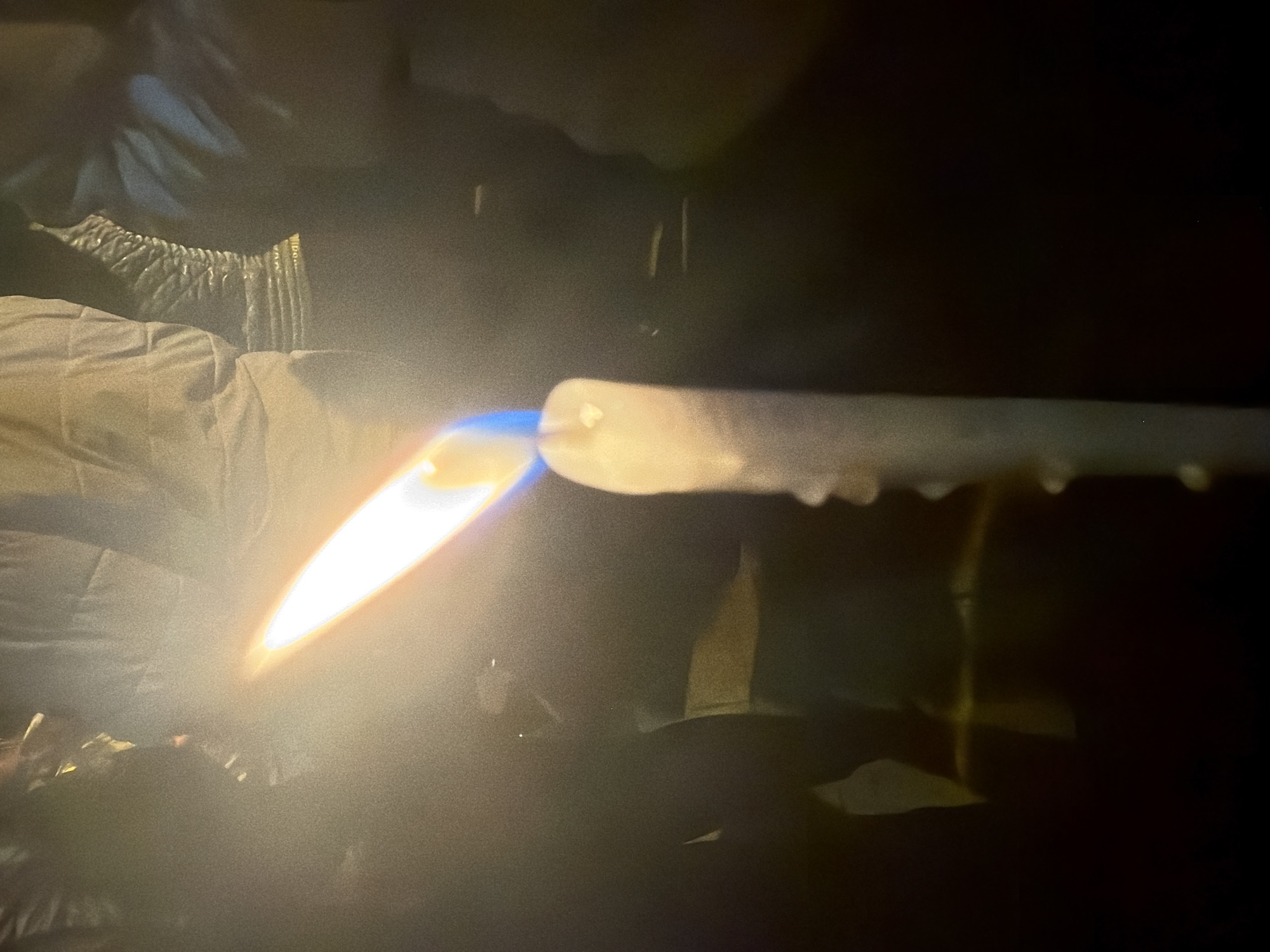 A person wearing what appears to be a quilted or textured sleeve is using a torch to heat a clear, cylindrical object. The torch emits a bright yellow and blue flame, and the heated object appears to be softening or melting, as evidenced by the drips on its side. The background is dark and obscured, but there are some faint shapes suggesting other objects or surroundings. The location is unspecified and not apparent from the image.