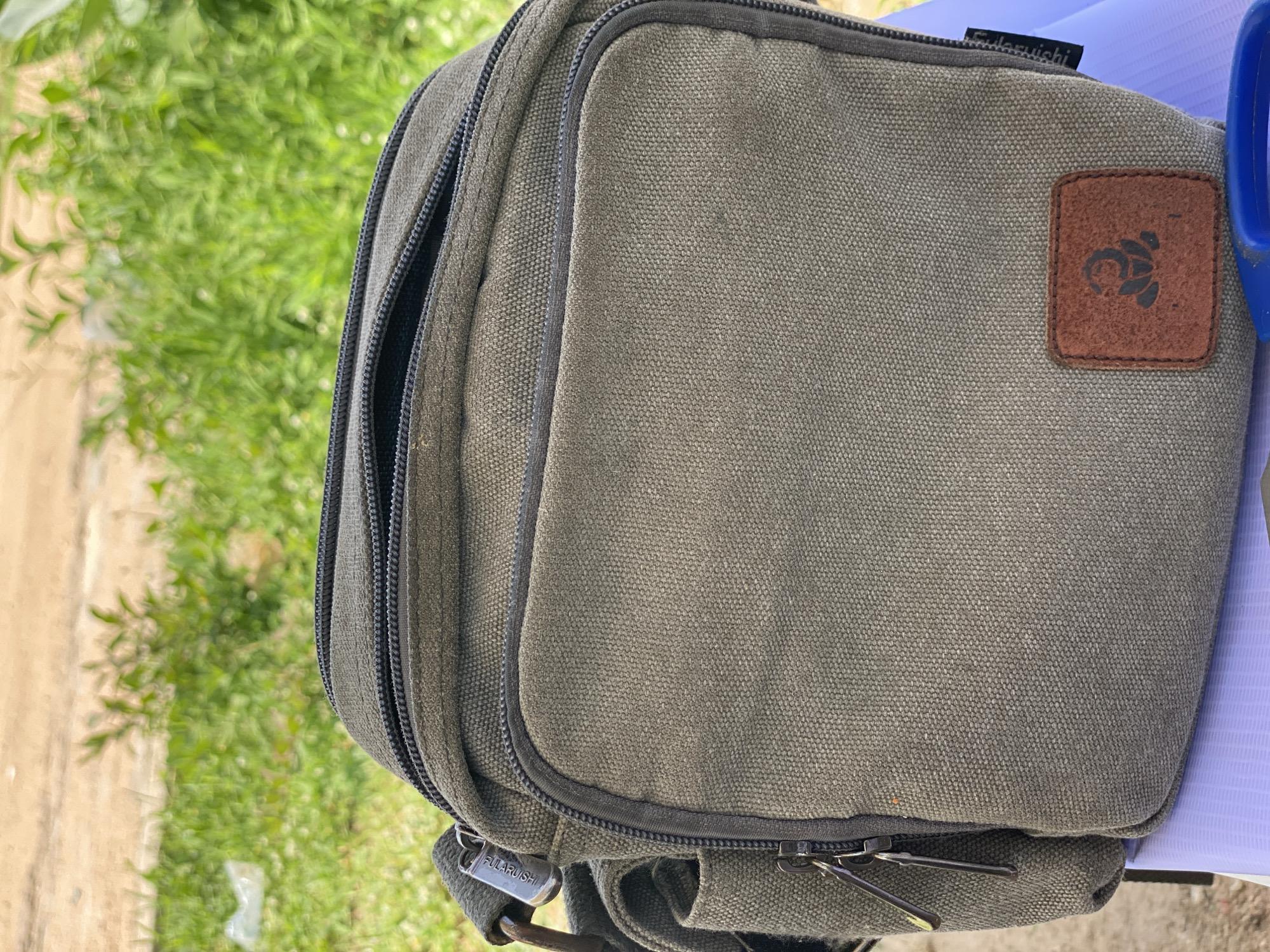 This is a close-up, medium-shot photograph of a green canvas bag with multiple zippers. The bag appears to be made of a durable material, possibly for outdoor use. It has a rectangular patch on the front with a dark emblem, possibly a logo, etched into it. There is also a small black tag attached to the bag with white text that reads "FULARUISHI". The bag is resting on a surface that appears to be a light purple chair or stool.
The background is out of focus but suggests an outdoor setting with green foliage and a dirt or gravel path. The lighting indicates it is daytime, and the overall ambiance is casual. The image is taken at an angle, with the bag dominating the frame. The focus is sharp on the bag, highlighting its texture and details. There is no indication of any people or specific activity taking place. The location context of Jalingo, Nigeria, does not provide any specific visual cues within the image itself, other than it being an outdoor scene.