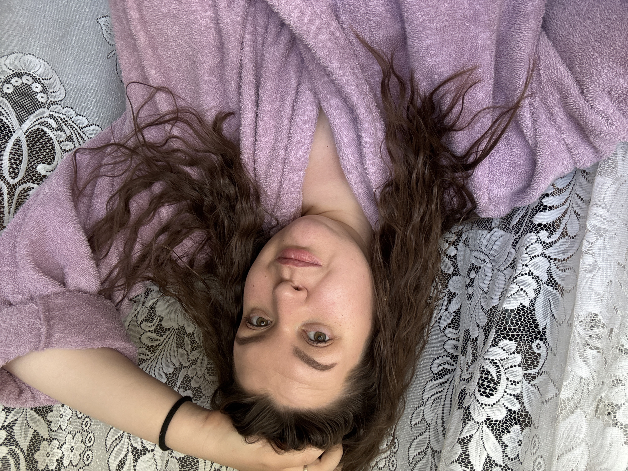 A woman is lying down, photographed from above. She is wearing a purple bathrobe. Her brown curly hair is spread around her head. She is resting her head on her hand, which has a black hair tie on the wrist. The background is a lacy white and grey patterned surface. It is impossible to determine the specific location in Марковский, Russia.