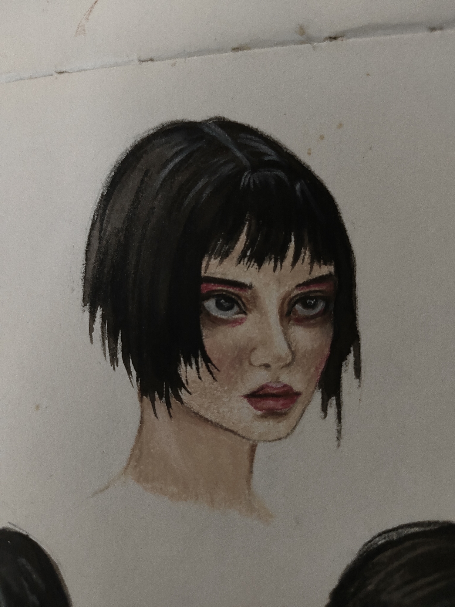 The image displays a close-up, angled photograph of a painted or drawn portrait on an off-white paper surface, located in the city of Unknown, Unknown. The central subject is an individual depicted from the upper chest, primarily focusing on the head and shoulders. The person has short, dark, possibly black, bob-cut hair with blunt, choppy bangs. The skin tone is light, and facial features include wide-set eyes with prominent dark circles or heavy shading underneath, augmented by distinct red eyeshadow or liner applied to both upper and lower eyelids. The irises appear a light, desaturated blue or grey. The nose is small and subtly contoured. The lips are closed, full, and painted a dark red or berry color. The overall expression is serious or intense. Below the subject's neck, the painting extends to show the beginning of the shoulders.
The paper background is uniformly light-colored, suggesting a plain backdrop for the portrait. Visible imperfections on the paper include several small, irregularly shaped brown speckles in the upper right quadrant. A faint horizontal crease or line is present across the top edge of the paper. In the bottom left corner of the image, a dark, rounded shape, resembling a portion of hair, is visible, suggesting either another incomplete figure or a continuation of the primary subject in a larger artwork not fully captured. The medium used for the artwork appears to be a form of watercolor, gouache, or acrylic, with visible brushstrokes and a slightly textured finish.