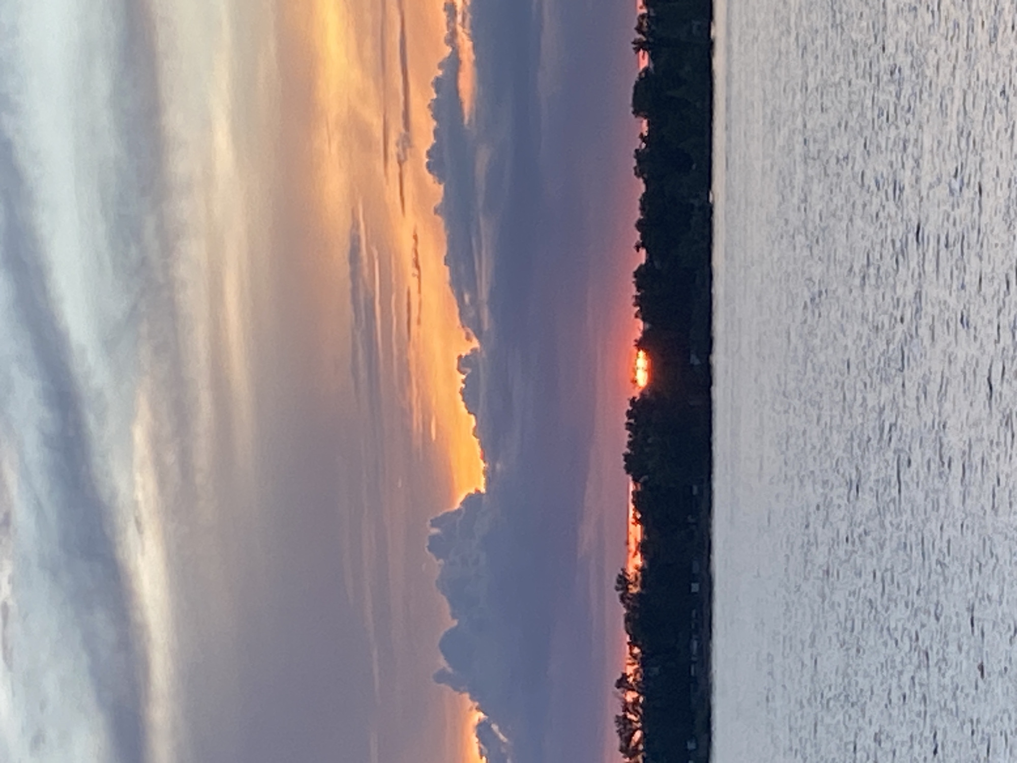 The image depicts a sunset over a body of water. The sky is filled with dramatic clouds, tinged with orange and purple hues, suggesting the sun is low on the horizon. A line of trees forms a dark silhouette along the far edge of the water, with hints of the setting sun breaking through the foliage at the horizon. The water itself appears rippled and reflects the muted colors of the sky. The scene evokes a sense of calm and the natural beauty of the transition from day to night. The time of day is clearly sunset. The weather appears clear, with scattered clouds. The location context is Novokuybyshevsk, Russia. There is no visible text in the image.