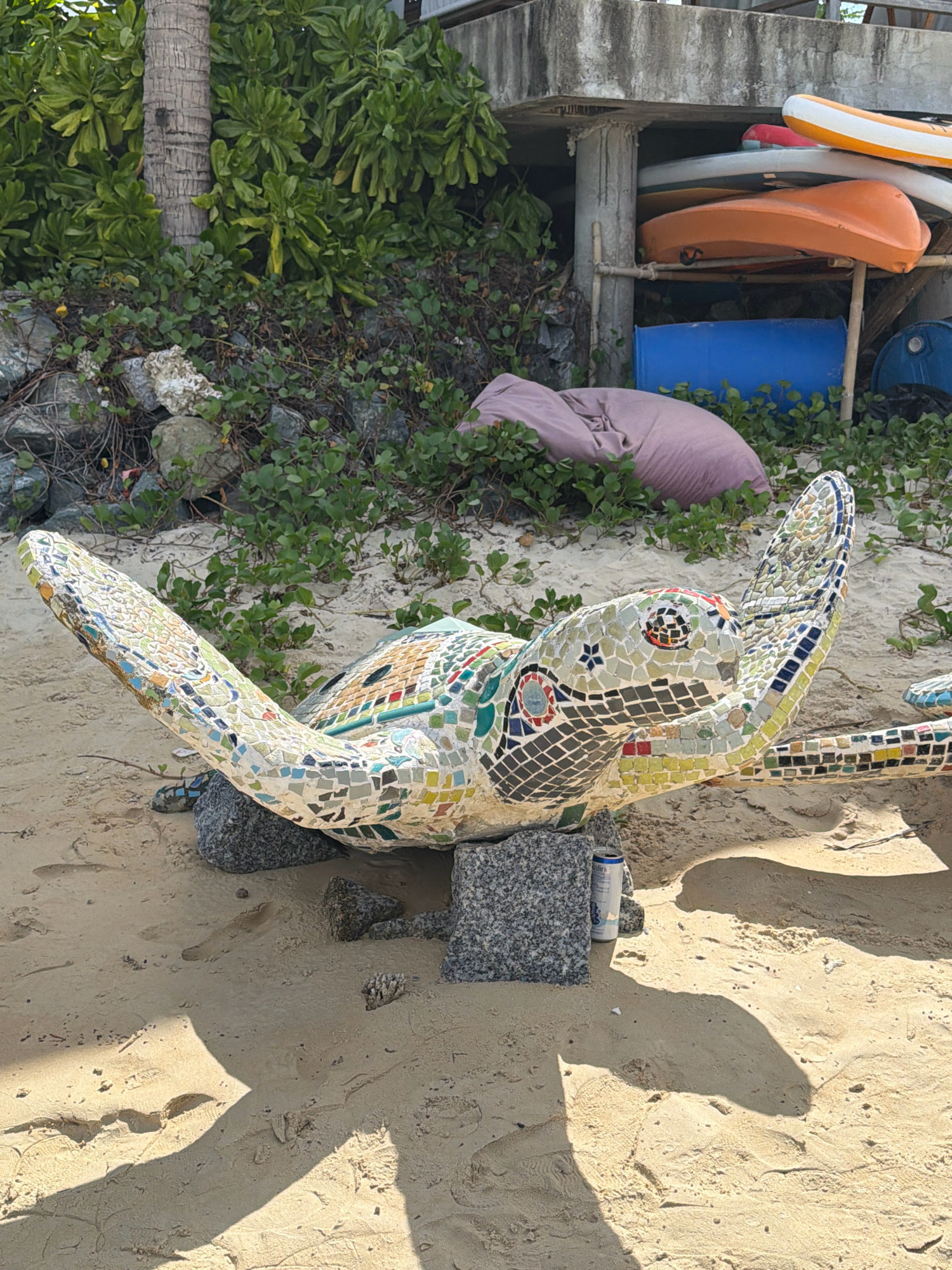 A mosaic sculpture of a sea turtle is positioned on a sandy beach in Nha Trang, Vietnam. The turtle sculpture, adorned with colorful tiles, is supported by several large rocks. In the background, lush green vegetation and palm trees are visible. Behind the vegetation, a concrete structure with stacked kayaks and barrels can be seen. A light purple beanbag chair is also present in the background. The sand shows footprints and some shadows cast by an unseen object. A can of drink is placed on the sand near one of the supporting rocks.