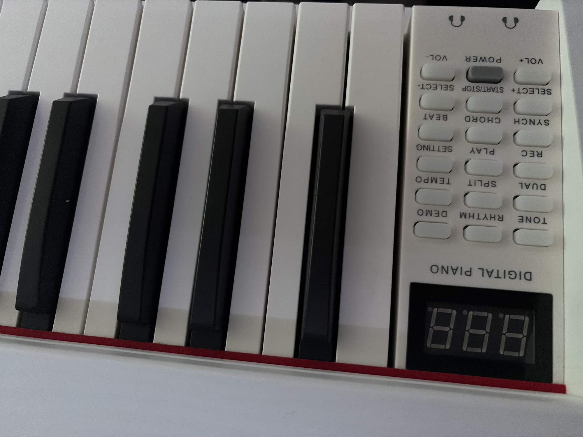 A close-up photograph shows a portion of a digital piano. The image features several white and black piano keys in the foreground. To the right of the keys is a control panel with various buttons labeled "VOL-", "POWER", "VOL+", "SELECT+ START/STOP SELECT-", "SYNCH CHORD BEAT", "REC PLAY SETTING", "DUAL SPLIT TEMPO", and "TONE RHYTHM DEMO." Below these buttons is a digital display showing "8.8.8.". The words "DIGITAL PIANO" are printed above the display. A thin red line runs horizontally along the bottom of the piano, visible between the keys and the white surface of the piano's body.