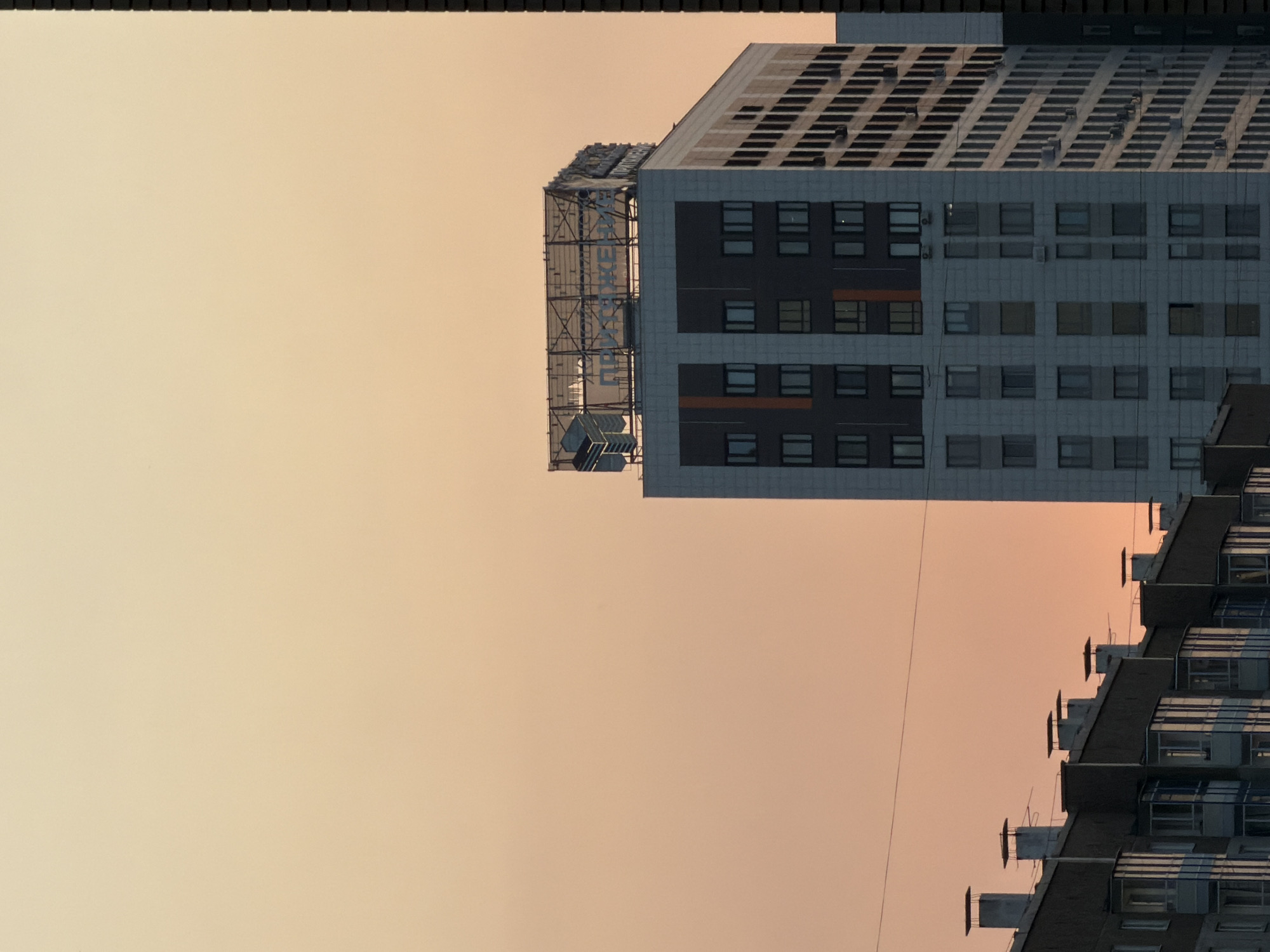 This image depicts an urban skyline at dusk, with a warm, gradient sky in shades of peach and yellow dominating the upper portion. In the mid-ground, a modern, multi-story building stands prominently. Its facade is a mix of light gray and darker gray panels, with rows of windows. The top of this building features a large, illuminated sign structure with what appears to be Cyrillic text and geometric graphics. To the left, the rooftops of several lower-rise buildings are visible, characterized by chimneys and balconies. The foreground reveals the lower sections of these buildings, showcasing balconies with blue tinted glass. To the right edge, a thin, dark vertical structure, likely another building, partially obscures the scene. The overall impression is one of a contemporary urban environment bathed in the soft light of sunset. This image was taken in the city of Krasnoyarsk, Russia, specifically within the residential complex "Pokrovsky" on Pokrovskaya Street.