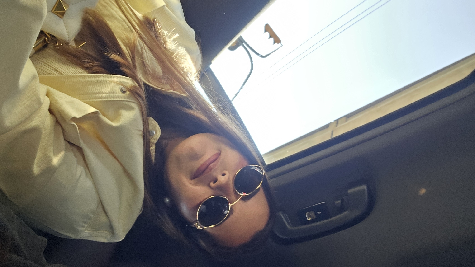 A young woman wearing sunglasses and a yellow jacket is pictured in what appears to be a car with a sunroof. The image is oriented with the woman's head tilted downwards towards the viewer. Sunlight streams through the sunroof. Overhead traffic lights are visible outside the sunroof. The location is presumed to be Mexico City, Mexico.