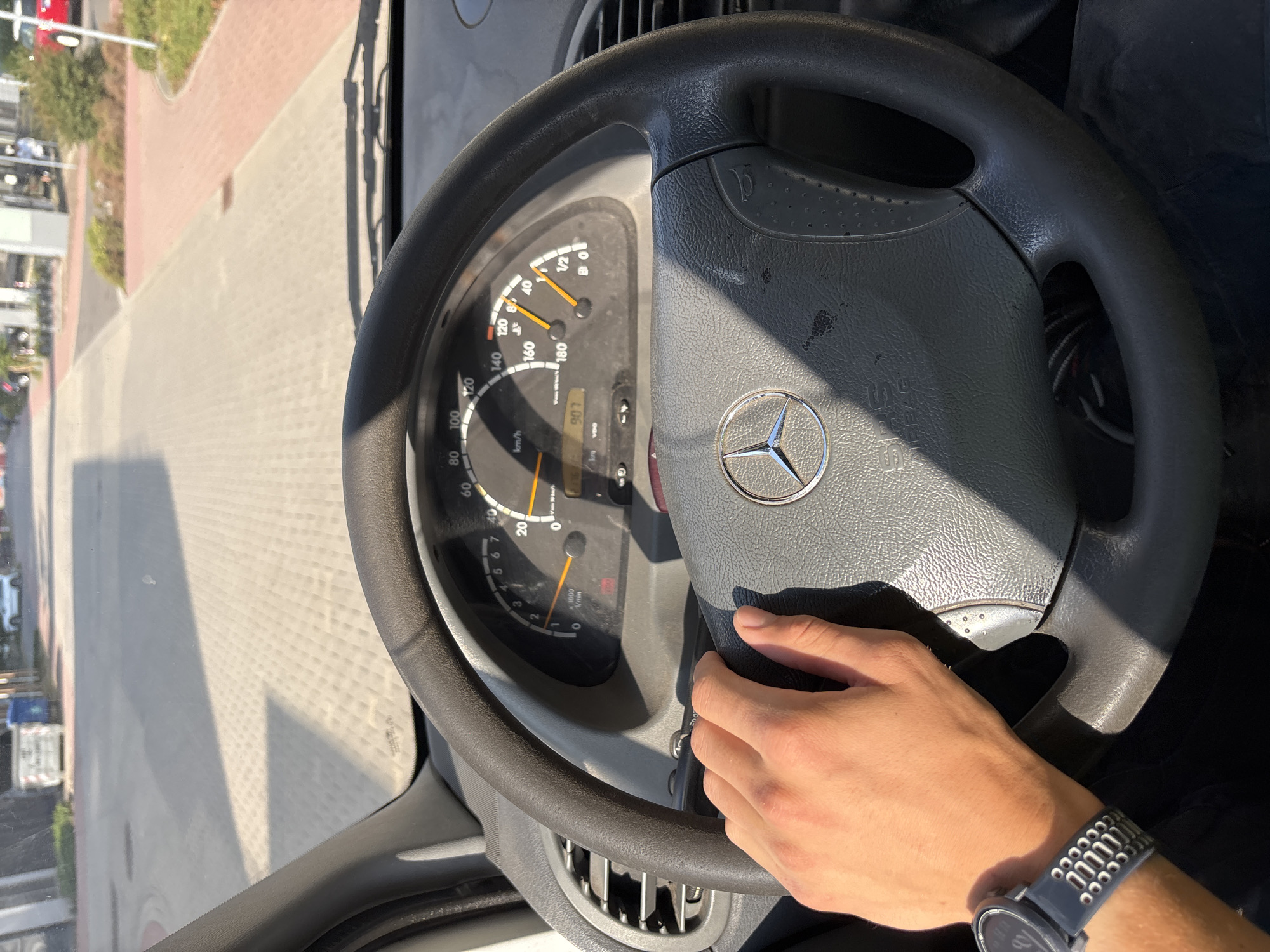 The image shows a first-person view from inside a vehicle, likely a van, as it travels down a street in Mannheim, Germany. The driver's left hand, wearing a dark wristwatch, is resting on the steering wheel. The steering wheel is dark grey and features the Mercedes-Benz logo in the center. The vehicle's dashboard is visible, with the speedometer indicating a speed of approximately 50 km/h. The road outside is paved with light-colored interlocking stones, and there are buildings and trees lining the street. The sun is casting long shadows, suggesting it is either morning or late afternoon. The overall scene depicts a typical urban driving environment in Mannheim on a sunny day.