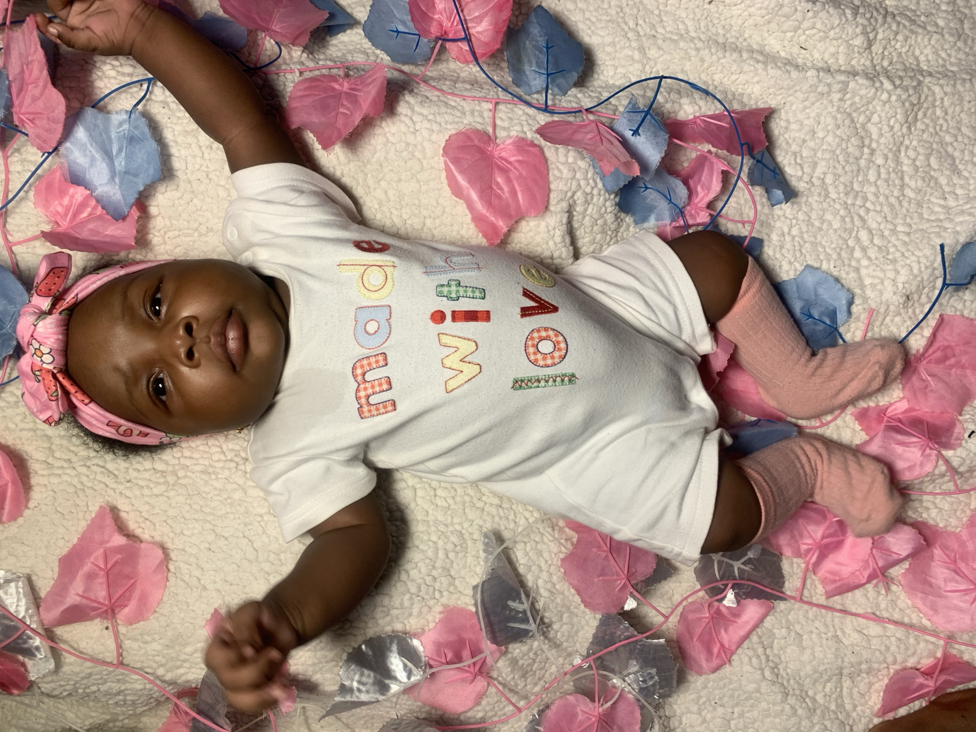 An overhead shot captures an infant lying on their back on a soft, light-colored blanket. The baby has dark skin, is wearing a white onesie, a pink headband with strawberry patterns, and light pink socks. The baby's head is tilted slightly, and their eyes are open, looking directly towards the viewer with a calm expression. A small gold stud earring is visible in the baby's left ear.
The white onesie has embroidered text that reads "made WITH LOVE." The word "made" is in a yellow and orange plaid pattern, "WITH" appears in a green plaid pattern with red outlines, and "LOVE" is in a red and orange checkered pattern.
Around the baby and on the blanket are decorative artificial vines with heart-shaped leaves. These leaves are predominantly pink and light blue, with some silver or grey ones also visible. The leaves are attached to thin, flexible stems, some pink and some blue, that crisscross around the baby. The setting appears to be indoors, likely a staged environment for a photograph, with the overall lighting suggesting an indoor or artificial light source. This scene is set in Rafin Bauna, Nigeria.