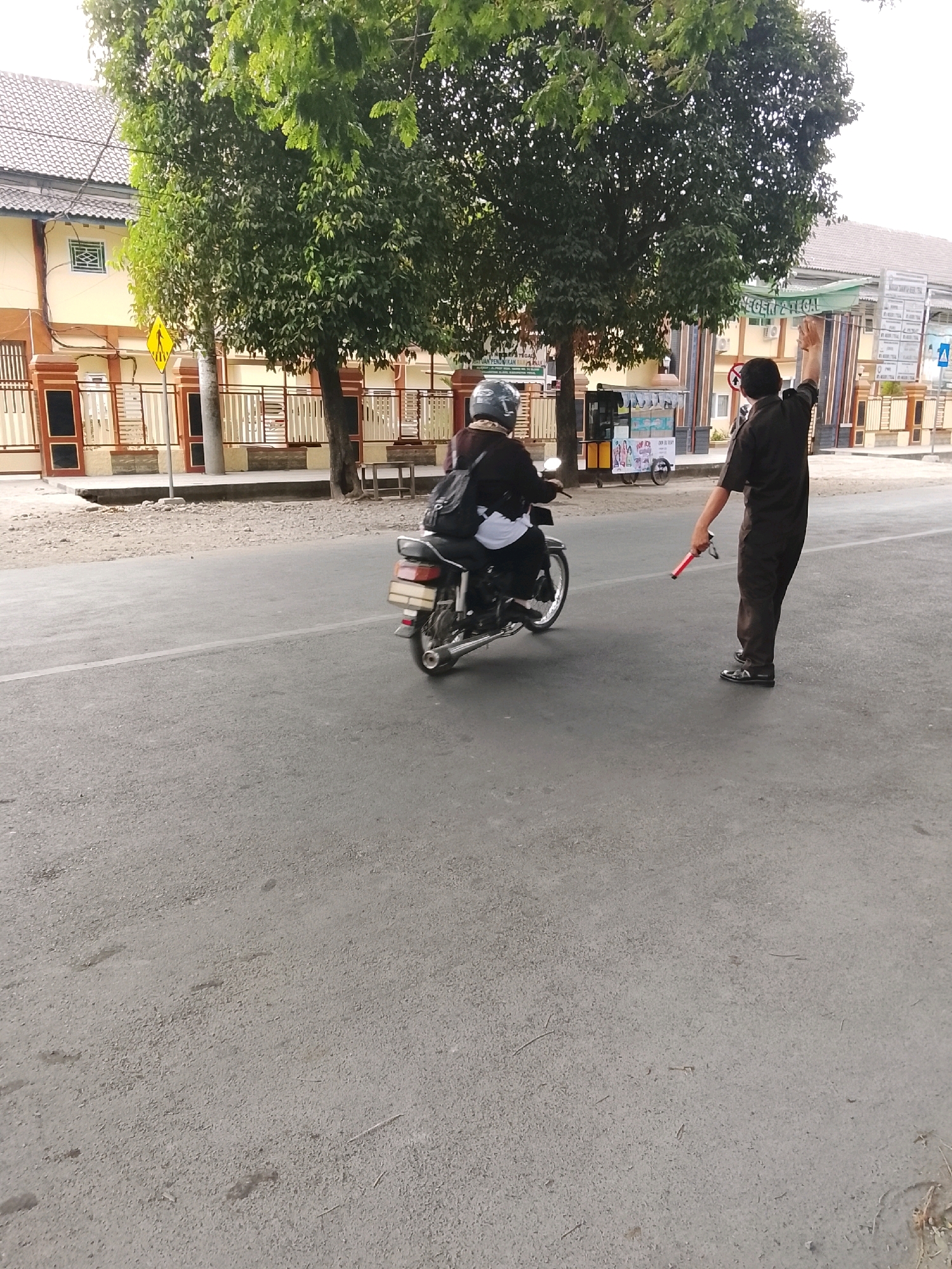 In Slawi, Indonesia, on a clear, sunny day, a man in dark attire, possibly a security guard or traffic enforcer, stands on the right side of a paved road, holding a red signaling baton in his right hand. His left arm is raised, signaling. To the left of the frame, a person wearing a helmet rides a motorcycle, facing away from the viewer. The motorcycle is black and has a luggage rack on the back. In the background, a building with a green awning that reads "KECAMATAN TEGAL" is visible. Various trees with lush green foliage line the side of the road. A yellow pedestrian crossing sign is also present on the left. The road surface is asphalt with a white line marking the lane.