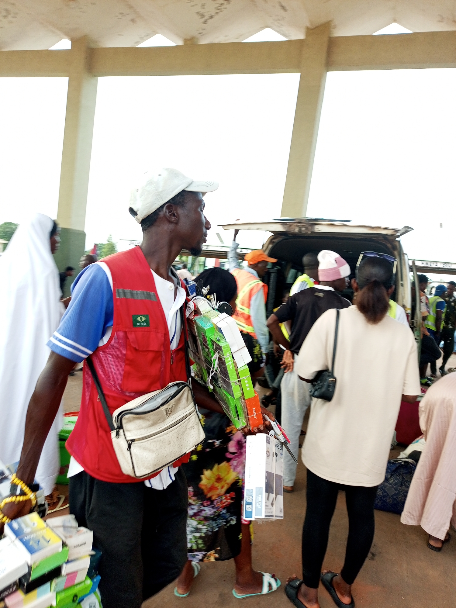 In the city of Gombe, Nigeria, a man wearing a white cap and a red vest over a blue and white striped shirt is seen carrying a stack of boxes. He has a light-colored shoulder bag with a black strap. To his left, a person in a white garment walks away from the camera. In the background, a van with its doors open is being loaded and unloaded by several individuals, some wearing high-visibility vests. The scene appears to be outdoors, possibly near a transportation hub or market.