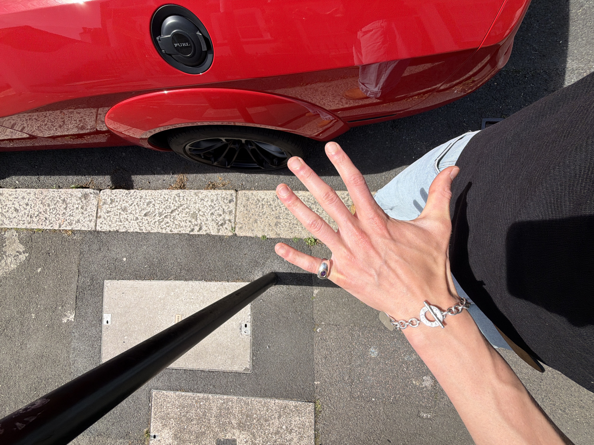 The image presents an eye-level view of a section of a street in London, United Kingdom. A red car is positioned in the upper left corner of the frame. The car has a fuel cap labeled "FUEL". The tires are black. The photograph includes a person’s arm and hand, adorned with a silver bracelet and a ring. The person is wearing jeans and a black shirt. A black pole is situated on the pavement. The pavement consists of asphalt and concrete tiles. The image is taken during the daytime in sunlight.