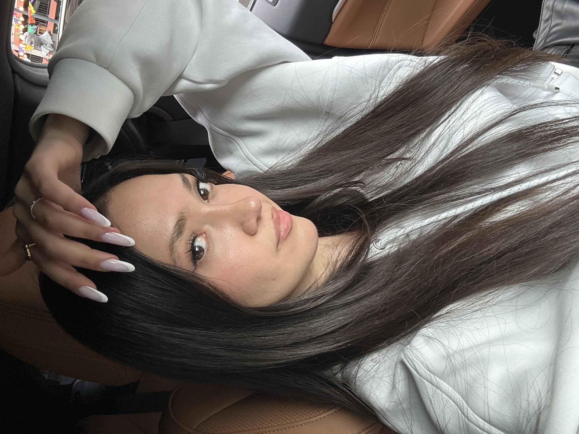 A woman lies on her side in a tan leather car seat, wearing a white hoodie. Her dark hair cascades across her face and the seat. Her hand, adorned with long, white-tipped nails, rests on her forehead. The background is blurry but suggests an urban environment seen through the car window. It is not possible to determine the exact location in Yerevan, Armenia, from this image alone.