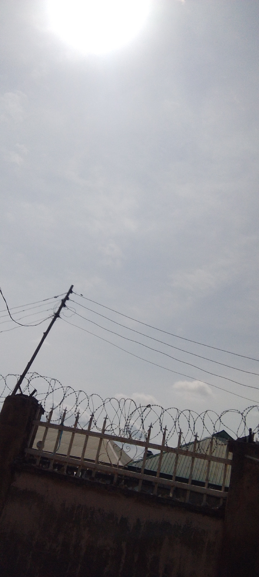 The image depicts an overcast sky with the sun partially visible, creating a bright halo in the upper left quadrant. A utility pole with multiple wires is angled across the frame from the lower left to the upper middle. In the foreground, a dark, textured wall runs along the bottom of the image. Above this wall, a light-colored metal fence is topped with barbed wire, creating a security barrier. Beyond the fence, the roofs of buildings are visible. The scene appears to be outdoors in an urban environment, though specific location details are unknown.