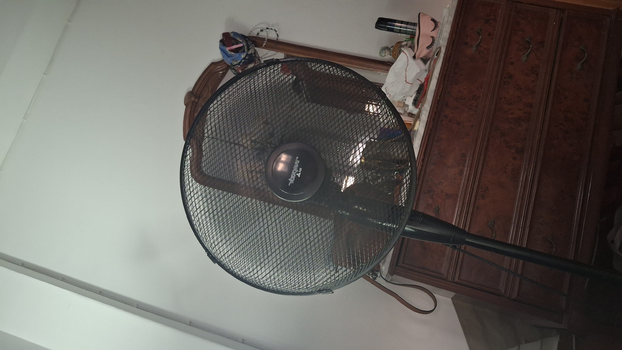 The image depicts an indoor scene, likely a bedroom, with a dark-colored floor fan positioned in the foreground. The fan's mesh grill is prominent, obscuring a portion of the background. Behind the fan, a wooden dresser with multiple drawers is visible. The dresser's surface is cluttered with various items, including a white bag, a makeup bag with eyelash designs, a bottle of hairspray, and other miscellaneous objects. A mirror with a wooden frame is also present, partially visible behind the fan. The walls appear to be white. The lighting suggests an artificial light source. The image does not contain any contextual information indicating a location in the city of Híjar, Spain.