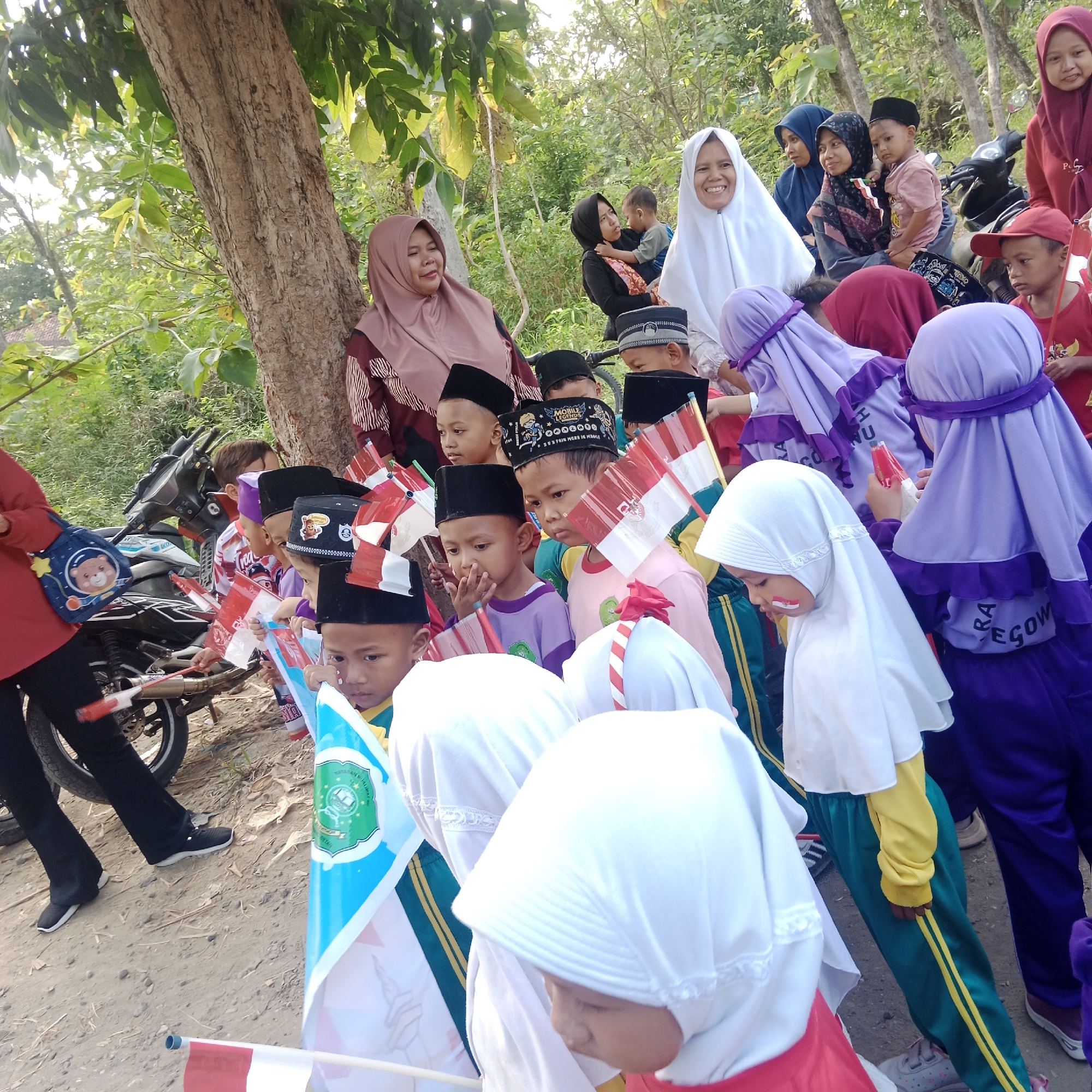In Tegowanu Kulon, Indonesia, a group of children, mostly boys, are gathered outdoors near a dirt road, holding small Indonesian flags. Many of the children are wearing traditional peci caps. Some of the children have red and white face paint, possibly representing the Indonesian flag. In the background, several adult women are also present, some wearing hijabs. A motorcycle is parked to the right of the frame. The environment appears to be a semi-rural area with trees and foliage. The overall scene suggests a community gathering or a local celebration.