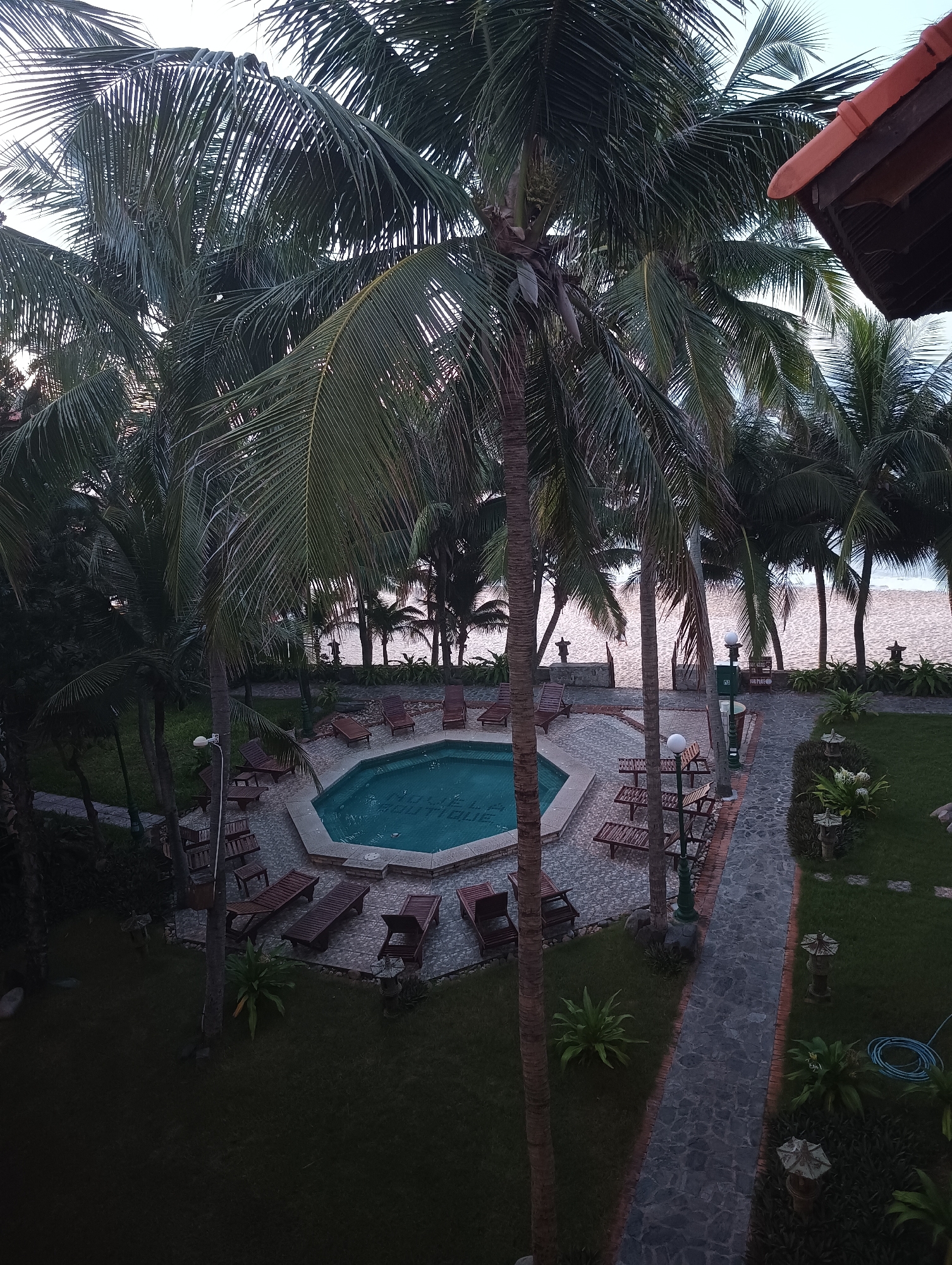 The image shows an aerial view of a resort swimming pool area in Phu Thuy Ward, Vietnam. Palm trees surround a hexagonal swimming pool, with lounge chairs and umbrellas set up on the stone patio. A pathway made of flagstone leads away from the pool area, bordered by manicured lawns and lush tropical foliage. In the background, a sandy beach meets the calm waters of the ocean. The sky is overcast, suggesting it is either early morning or late afternoon. The resort appears to be a tranquil and relaxing destination.