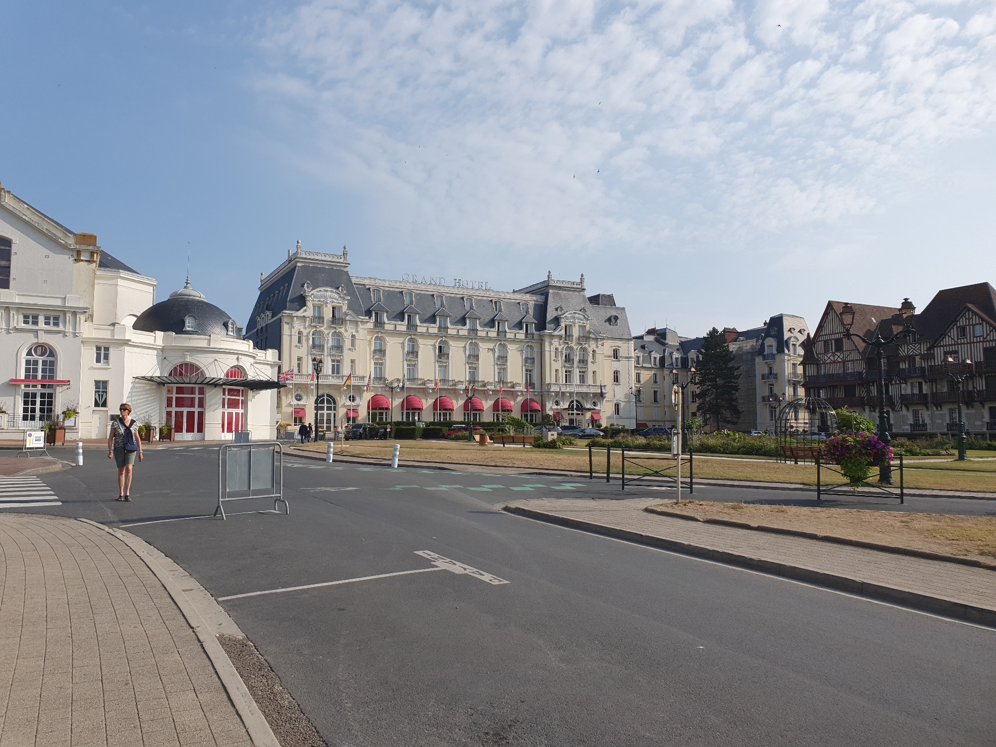 A street view of the Grand Hotel in Cabourg, France, under a partly cloudy sky. A woman wearing a patterned shirt and shorts, with a blue backpack, walks across a crosswalk on the left side of the image. In the background, the Grand Hotel, a large, ornate building with a grey mansard roof and cream-colored facade, is visible. Red awnings cover many of the windows. To the right of the hotel are several half-timbered buildings. A road runs through the foreground, with a sidewalk and a grassy area to the right. Several streetlights and decorative metal fences are also present. Cars are parked in front of the hotel.