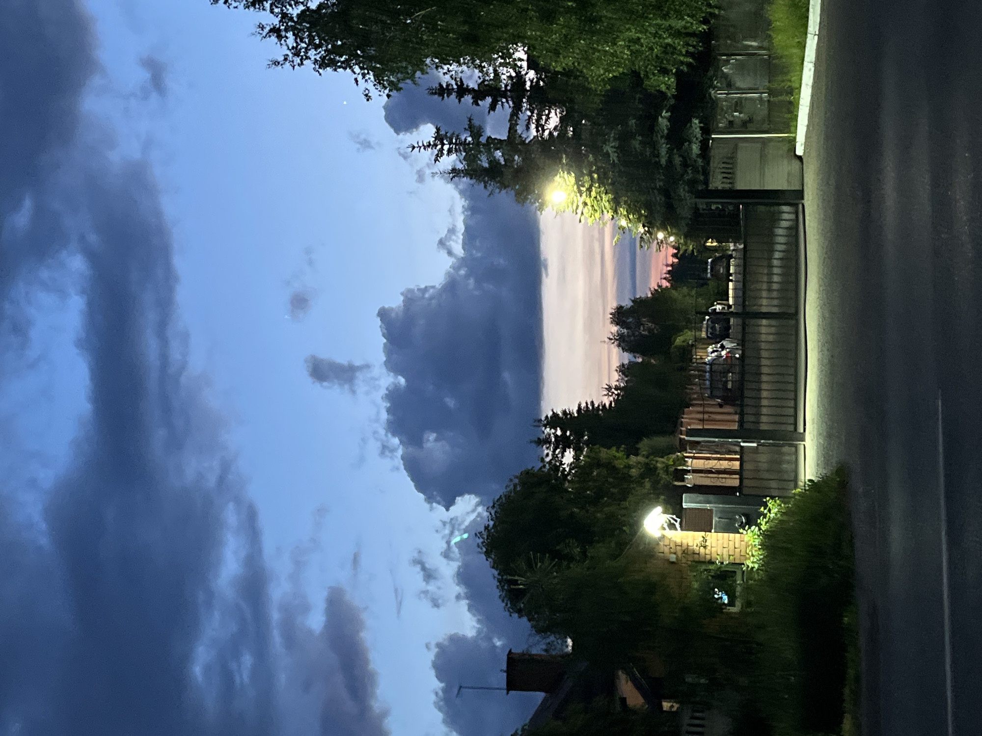 This image captures an outdoor scene at dusk or early night in what appears to be a residential or semi-rural area in Orekhovka, Russia. The sky dominates the upper portion of the frame, showcasing a dramatic blend of deep blues and dark, voluminous grey clouds on the left. Towards the right horizon, the clouds thin, revealing a soft gradient of pink and orange hues from a fading sunset.
Lush green trees line the mid-ground, some appearing as dark silhouettes against the twilight sky, while others are brightly illuminated by artificial lights. Several glowing streetlights or outdoor lamps cast warm, yellowish light, highlighting sections of foliage and structures. A dark paved road or path runs along the bottom, with a faint white line marking its edge.
In the mid-ground, glimpses of buildings or houses are visible, characterized by rooftops and a chimney. A prominent dark metal gate stands on the right, behind which the faint outlines of parked vehicles can be seen. The scene is devoid of people or discernible activity, conveying a quiet and transitional atmosphere. No visible text is present.