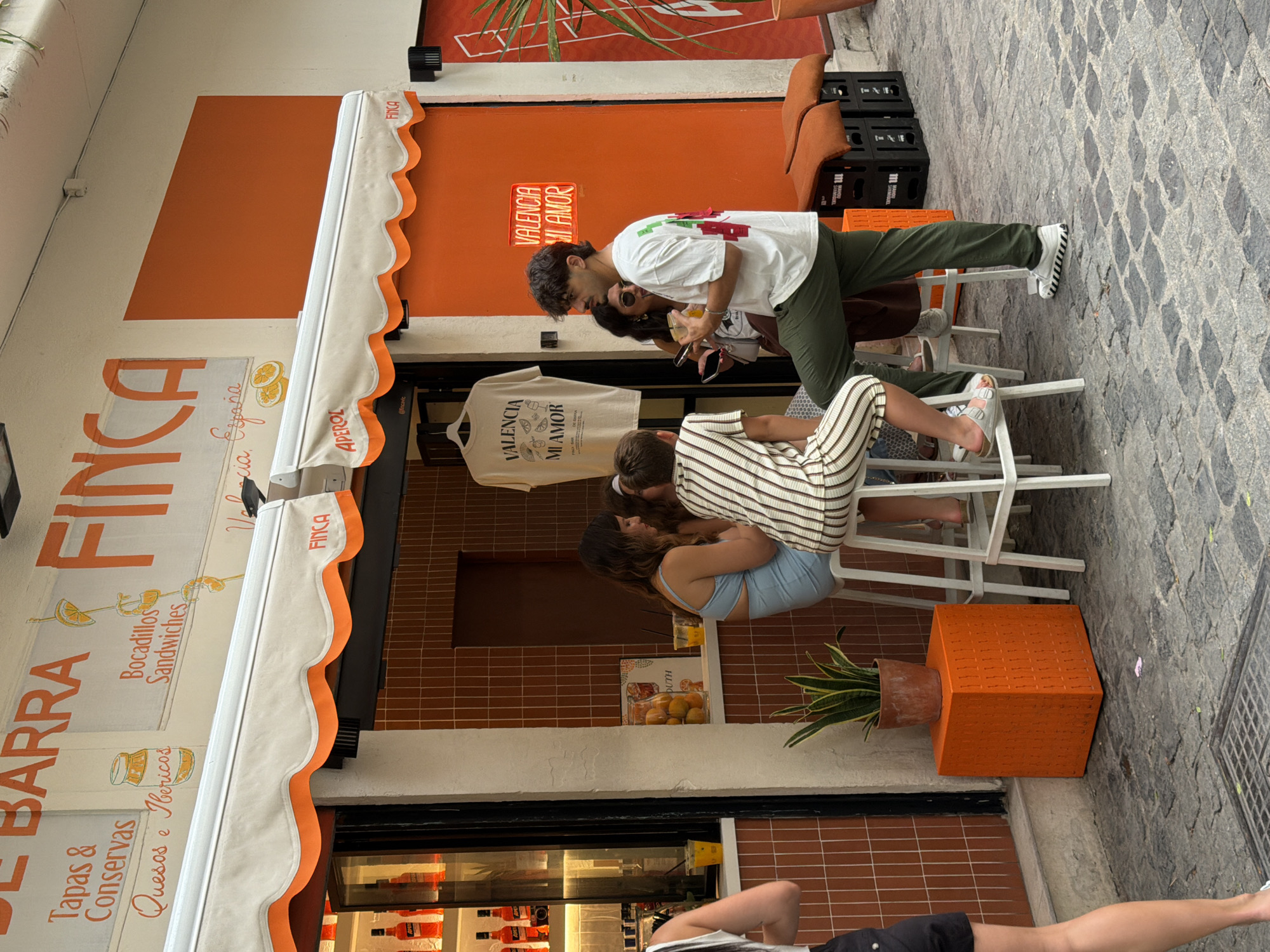 The image shows a street scene in front of a vibrant orange and white establishment identified as "FINCA" and featuring the phrase "VALENCIA MI AMOR" in neon lights. A white awning with orange trim extends over the entrance. Two women and a man are visible. One woman, wearing a light blue outfit, sits on a white stool at a counter. Another woman, dressed in a striped outfit, is also seated at the counter. A man in a white t-shirt and green pants stands facing the seated individuals, holding a drink. The ground is paved with cobblestones. Various objects are present, including a potted plant on an orange cube, a display of bottled drinks inside the establishment, and stacked crates with orange cushions outside. The overall aesthetic suggests a casual, outdoor cafe or bar setting.