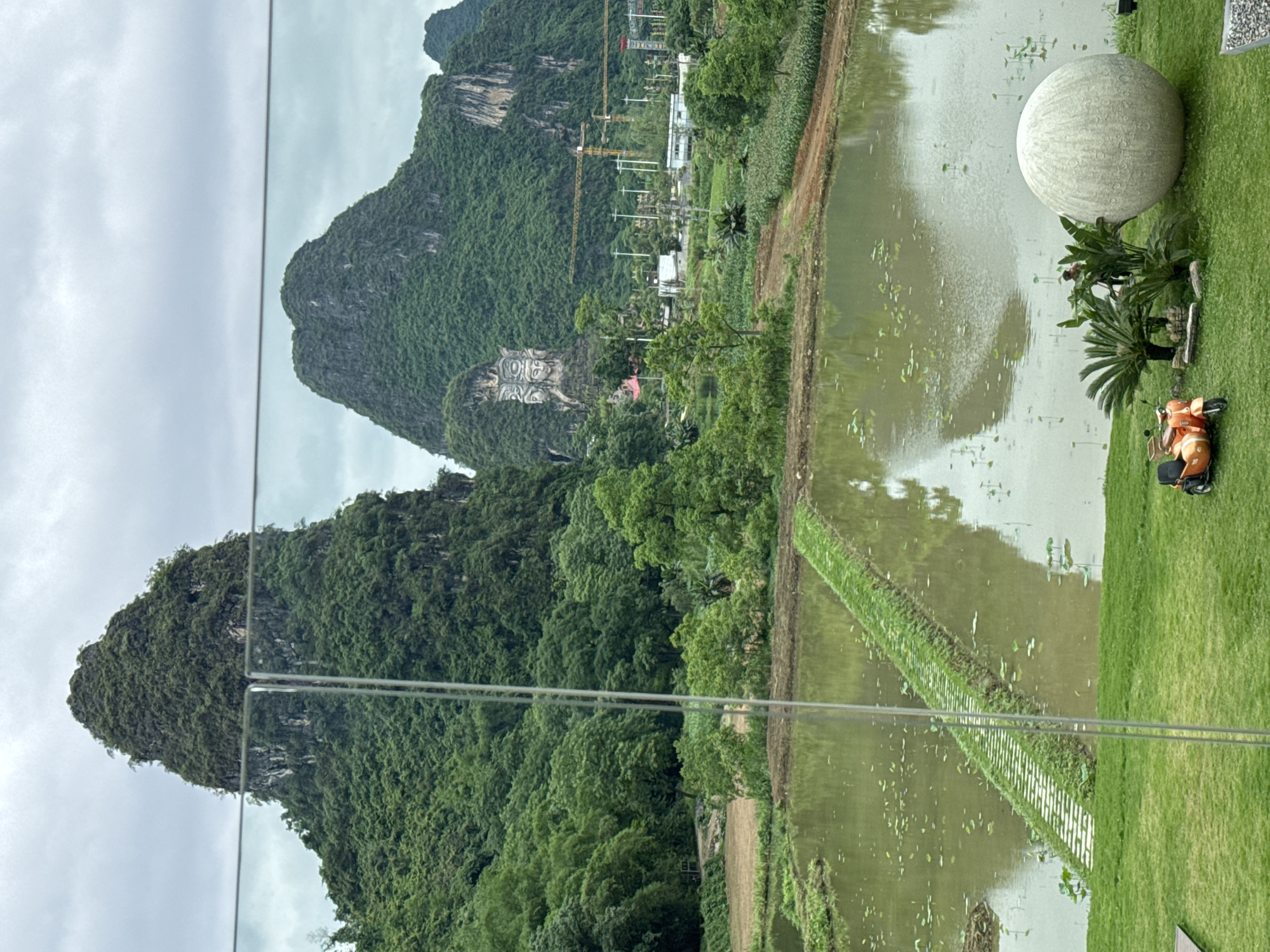 The photograph depicts a landscape in Guilin, China, characterized by verdant karst mountains. In the background, one of the mountains features a carved image. In the foreground, there is a body of water that appears to be a pond or lake, reflecting the sky and surrounding foliage. Alongside the water, there is a grassy area with a large spherical stone or art installation. A small orange scooter is parked on the grass near the art installation. There are visible power lines running across the lower portion of the frame.