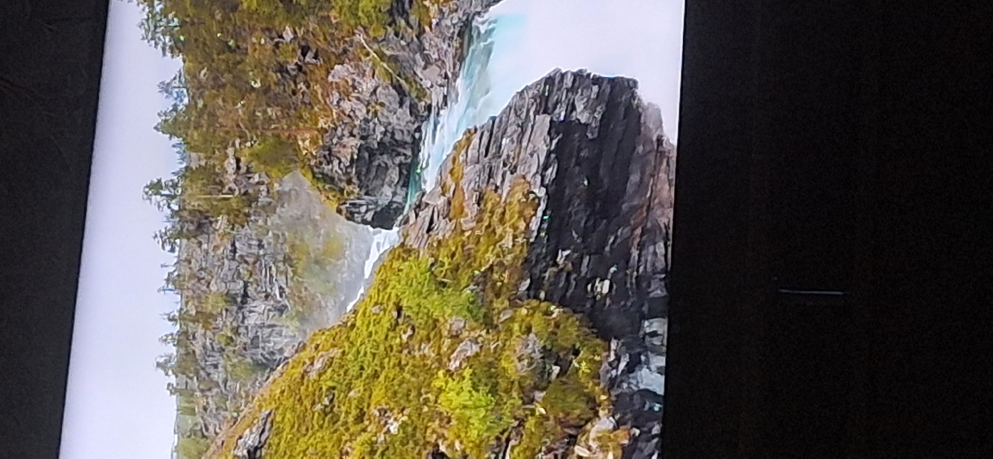 The image shows a scenic view of a river flowing through a rocky gorge. The landscape is characterized by steep, moss-covered slopes and rugged rock formations. Pine trees are scattered along the tops of the cliffs. The river itself is a bright, turquoise color and appears to be moving rapidly, with white water rapids visible. The water reflects the sky and surrounding landscape. There is no indication that this image is located in Bila Tserkva, Ukraine, or any other specific city. The image is a landscape photograph, possibly of a natural park or wilderness area.