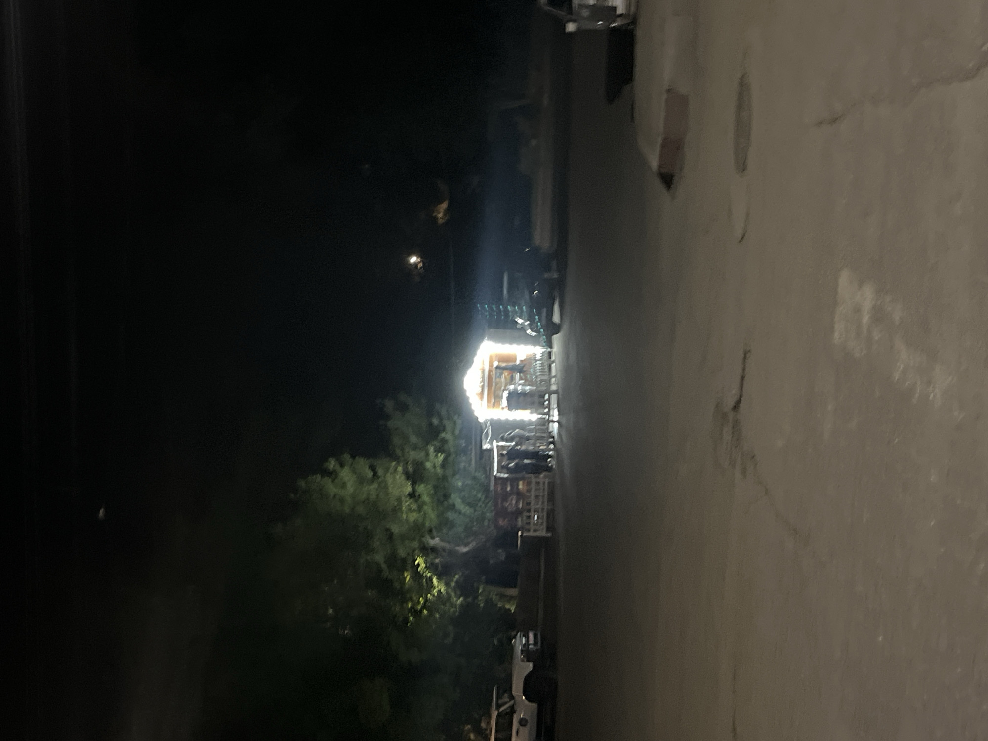 The image shows a dimly lit outdoor scene at night. In the background, there is a brightly lit roadside stall or kiosk with several people standing near it. A large tree with green foliage is visible behind the stall. To the left of the stall, a white SUV is parked. Further to the right, another dark-colored vehicle is visible, along with part of what appears to be another vehicle in the foreground on the right edge of the image. The ground in the foreground is paved and shows some cracks and circular manhole covers. The overall atmosphere is that of a street scene at night, possibly a market or a gathering place.