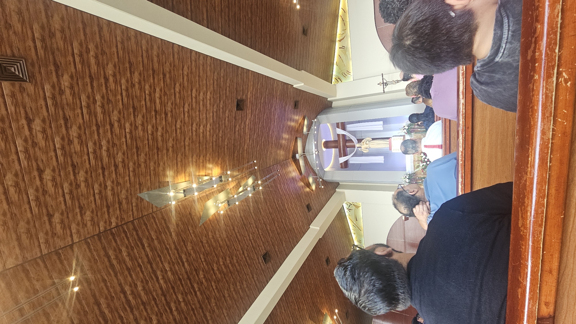 A church interior in San Bernardino Tlaxcalancingo, Mexico. A high, brown, wood-paneled ceiling is lit by modern chandeliers, with white trim along the ceiling and vertical supports. People are seated in wooden pews, facing an altar with a cross and statue. Visible details include hair color, shirt colors (blue, black, purple, gray), and facial profiles. The scene suggests a religious service in progress.