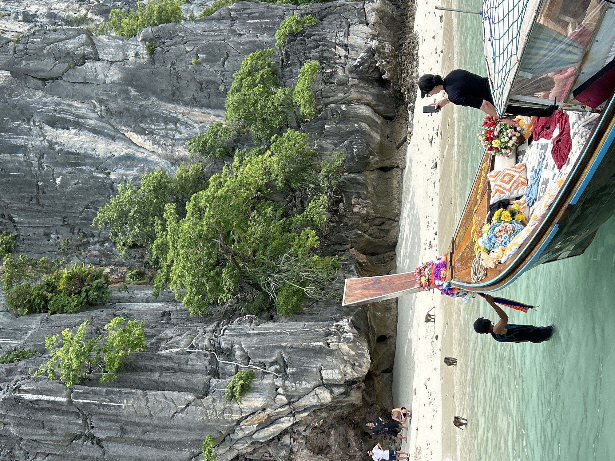 In the city of Ton Sai, Thailand, on a bright day, a long-tail boat is moored in clear, turquoise water near a sandy beach and a towering, sheer grey limestone cliff face overgrown with lush green vegetation. Several monkeys are scattered across the sand and in the shallow water near the boat. A person, partially submerged in the water by the boat, is holding a colorful sash. Another person, wearing a black shirt and cap, is seated on the boat, holding up a smartphone as if taking a picture. The boat is adorned with vibrant floral arrangements and decorative pillows. In the background, on the beach, three individuals are visible standing near the base of the cliff.