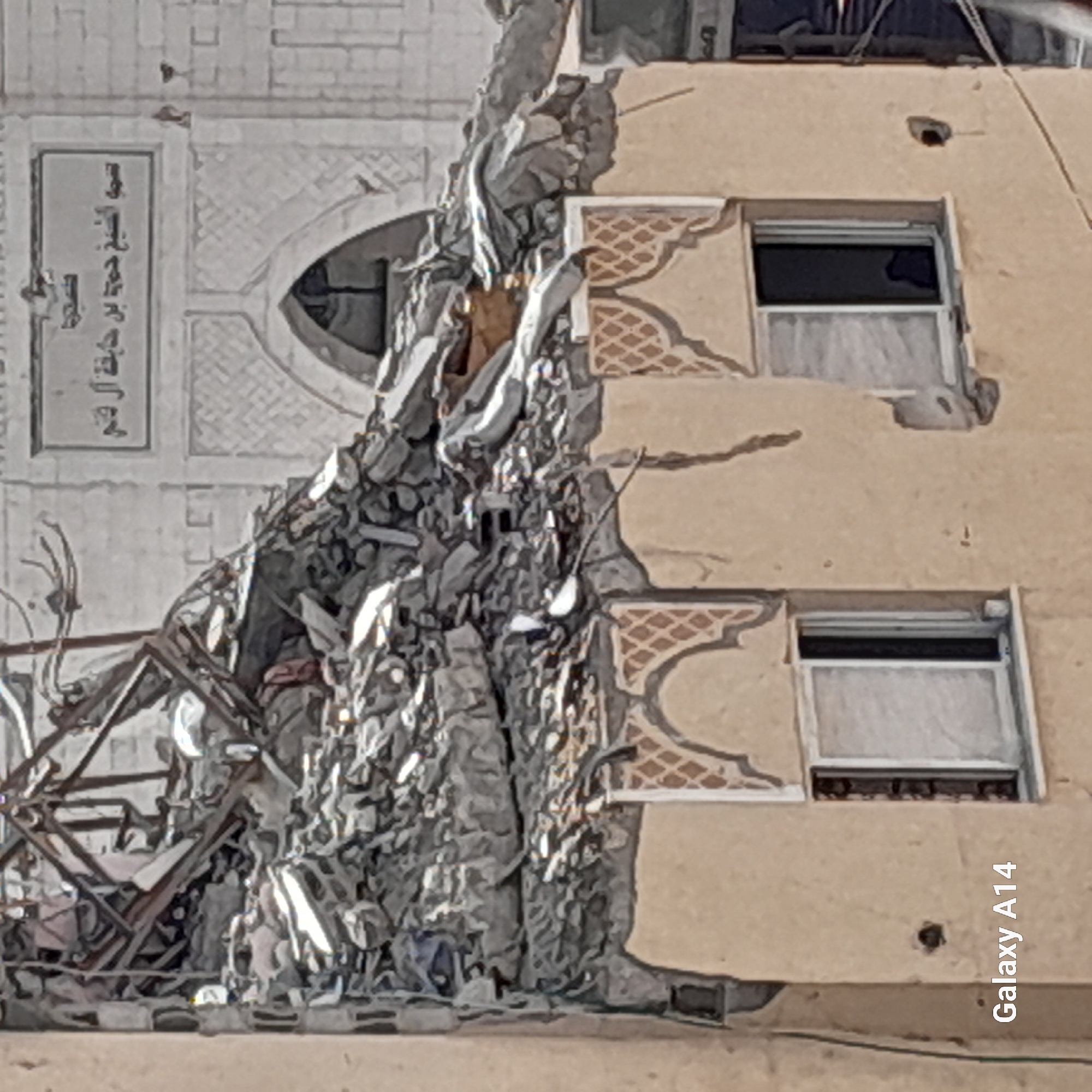 The image shows the exterior of a damaged building in Khan Yunis, Palestinian Territories. The top portion of the building is heavily damaged, with rubble and debris indicating a structural collapse. There are visible metal beams and concrete fragments. The lower part of the building features a tan-colored facade with decorative patterns above two windows. The windows are rectangular with white frames, and the glass panes are visible, with one appearing to be broken. The overall scene suggests destruction and likely an impact or bombing event. The text "Galaxy A14" is overlaid on the bottom left corner of the image, indicating the device used to capture the photograph.