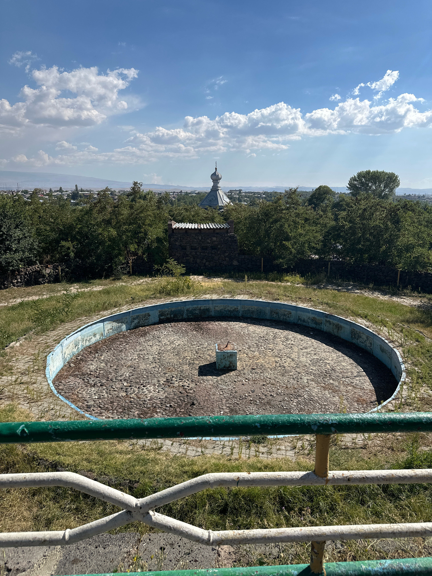 This is a photograph of an empty, circular, concrete basin with a low blue rim, situated in an outdoor environment. The basin is filled with small, grey, rounded stones. In the center of the basin, a small, rectangular, blue concrete pedestal stands. The basin is surrounded by dry grass and scattered weeds. A railing, made of green and white metal bars, is visible in the foreground. In the background, beyond a line of trees, a town or city is visible with buildings, and a distinctive domed structure with a spire, likely a church, rises above the rooftops. The sky is blue with scattered white clouds. This scene is located in Gyumri, Armenia, likely at the abandoned outdoor swimming pool of the Gyumri Water Park.